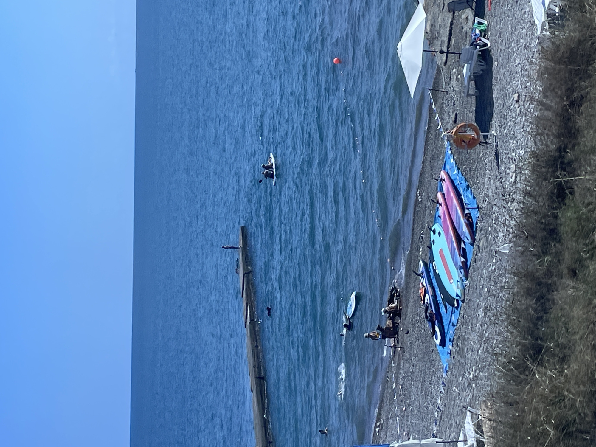 The image depicts a sunny day at a beach in Sochi, Russia. The foreground shows a pebbly shore with several surfboards laid out on blue tarps. Further back, people are visible on the beach and in the water. Some individuals are seated on the shore, while others are swimming or engaged in water activities. A long concrete pier extends into the blue sea, with a lone figure standing at its end. Further out in the water, two people are paddling a board, and an orange buoy floats nearby. The sea is calm and stretches to the horizon under a clear blue sky. The environment is characteristic of a coastal resort area.