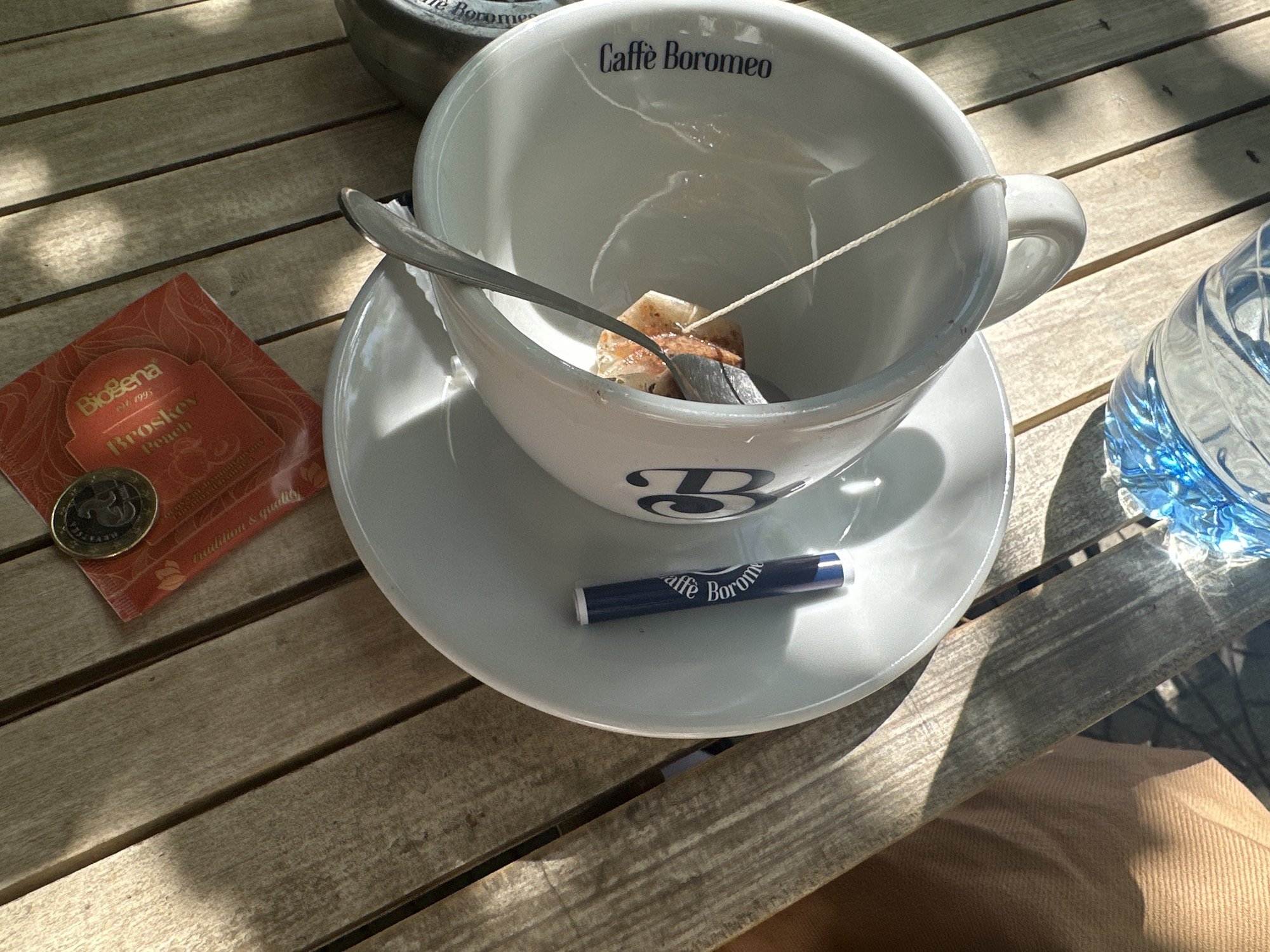 On a wooden planked table, presumably outdoors, sits a white ceramic teacup with the words "Caffè Boromeo" printed on it, a saucer, a tea bag, a spoon, a sugar packet with "Caffè Boromeo" printed on it, and a packet of "BioSena Broskov Peach" tea with a coin on top. A bottle of water is on the right. Part of a person's khaki-colored clothing is visible in the lower right corner. The location appears to be a café in Sofia, Bulgaria, based on the "Caffè Boromeo" branding.