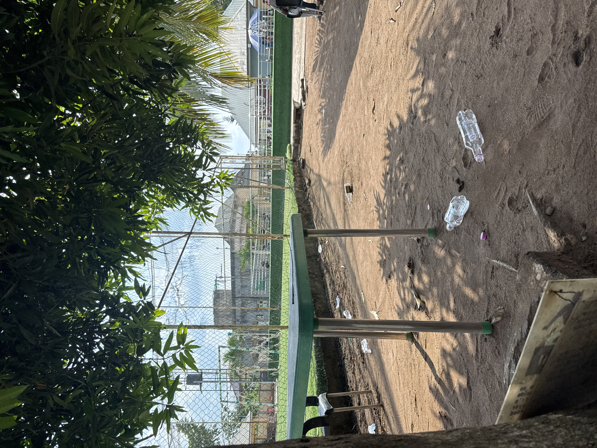 The image shows an outdoor scene in Ihiagwa, Nigeria. A green table stands in front of a chain-link fence. Beyond the fence, there's a field or sports area with structures suggesting a stadium or event space. In the foreground, the ground is sandy with litter, including plastic water bottles. Trees and foliage partially obscure the view on the left. The photograph is rotated. The lighting suggests a sunny day. Some people can be seen inside the stadium beyond the fence. A sign board is on the right side.