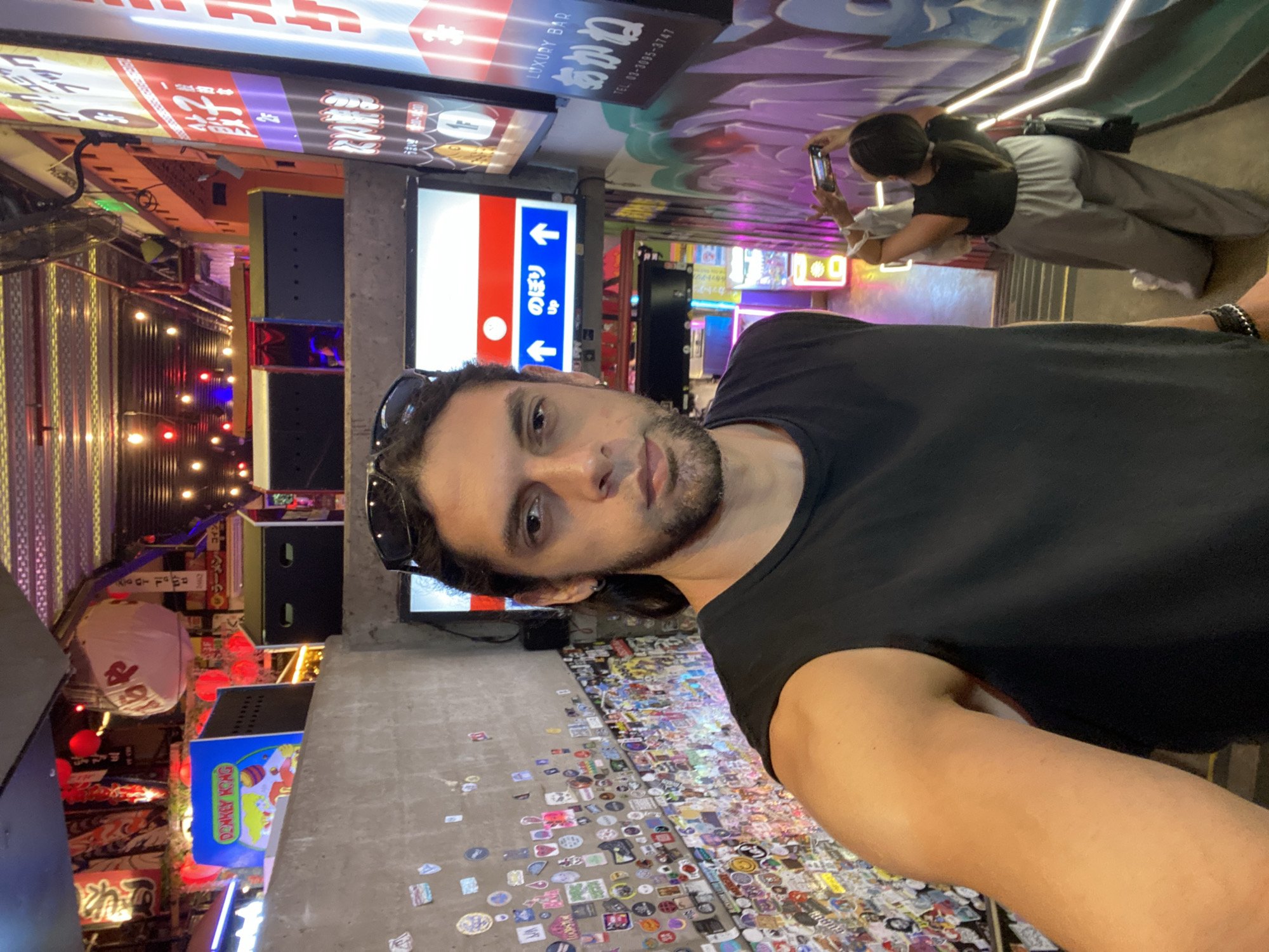 A man in a black t-shirt stands in an alleyway, taking a selfie. The alleyway is decorated with neon signs and stickers. In the background, a person is taking a picture with a smartphone. Signs in Japanese are visible.
