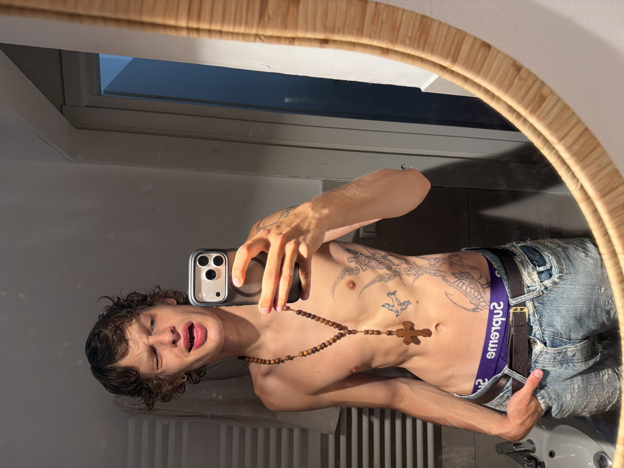 A young, slender, shirtless man with brown curly hair is taking a mirror selfie in a bathroom. He's wearing blue ripped jeans with a brown belt and purple underwear with "Supreme" written on it. He is holding a smartphone in his right hand. The man has tattoos on his torso, chest, and arms. He's also wearing a wooden rosary with a cross. The mirror has a round wicker frame. Reflected in the mirror are a bathroom sink, a radiator, a window, and a white wall. The location cannot be pinpointed to Milan, Italy from the available image.