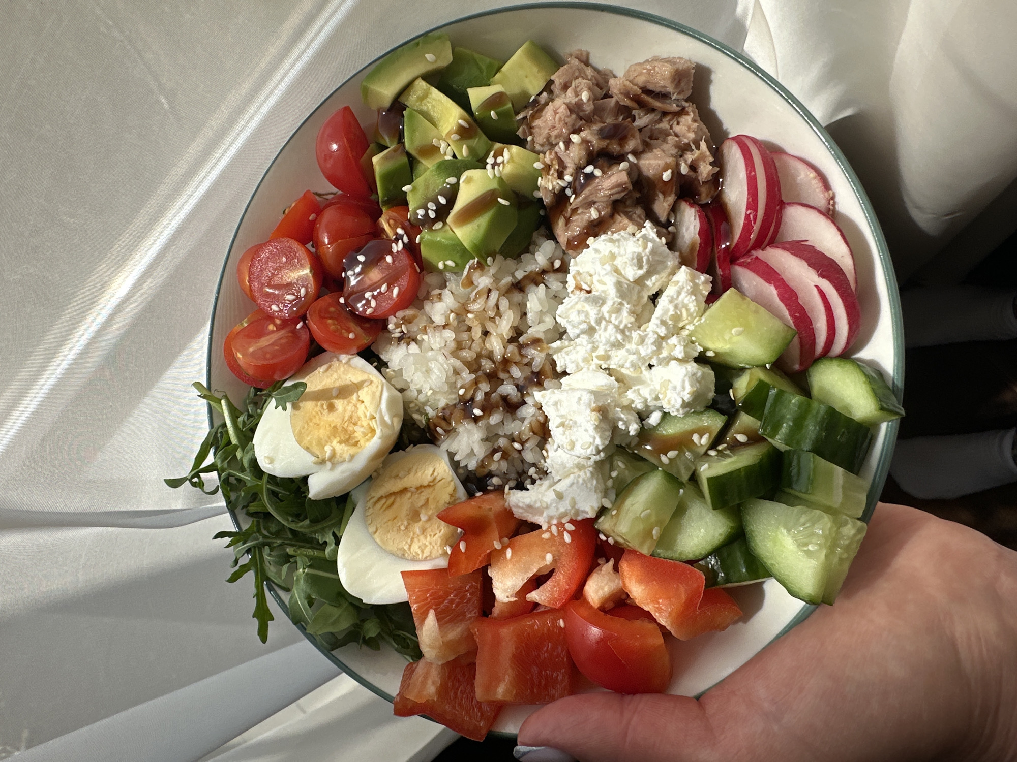 A well-lit close-up shot depicts a vibrant salad bowl held in someone's hand. The bowl contains an assortment of ingredients artfully arranged: sliced avocado, halved cherry tomatoes, cooked rice with dark sauce, a dollop of white cheese (possibly feta or cottage cheese), sliced radishes, diced cucumber, diced red bell pepper, hard-boiled egg halves, and arugula. The ingredients appear to be drizzled with a dark sauce and sprinkled with sesame seeds. The bowl has a white interior with a thin green rim. The hand holding the bowl has pale skin and a light gray nail polish. The background is a soft white fabric. There is no indication that this image was taken in Chaikovsky, Russia.