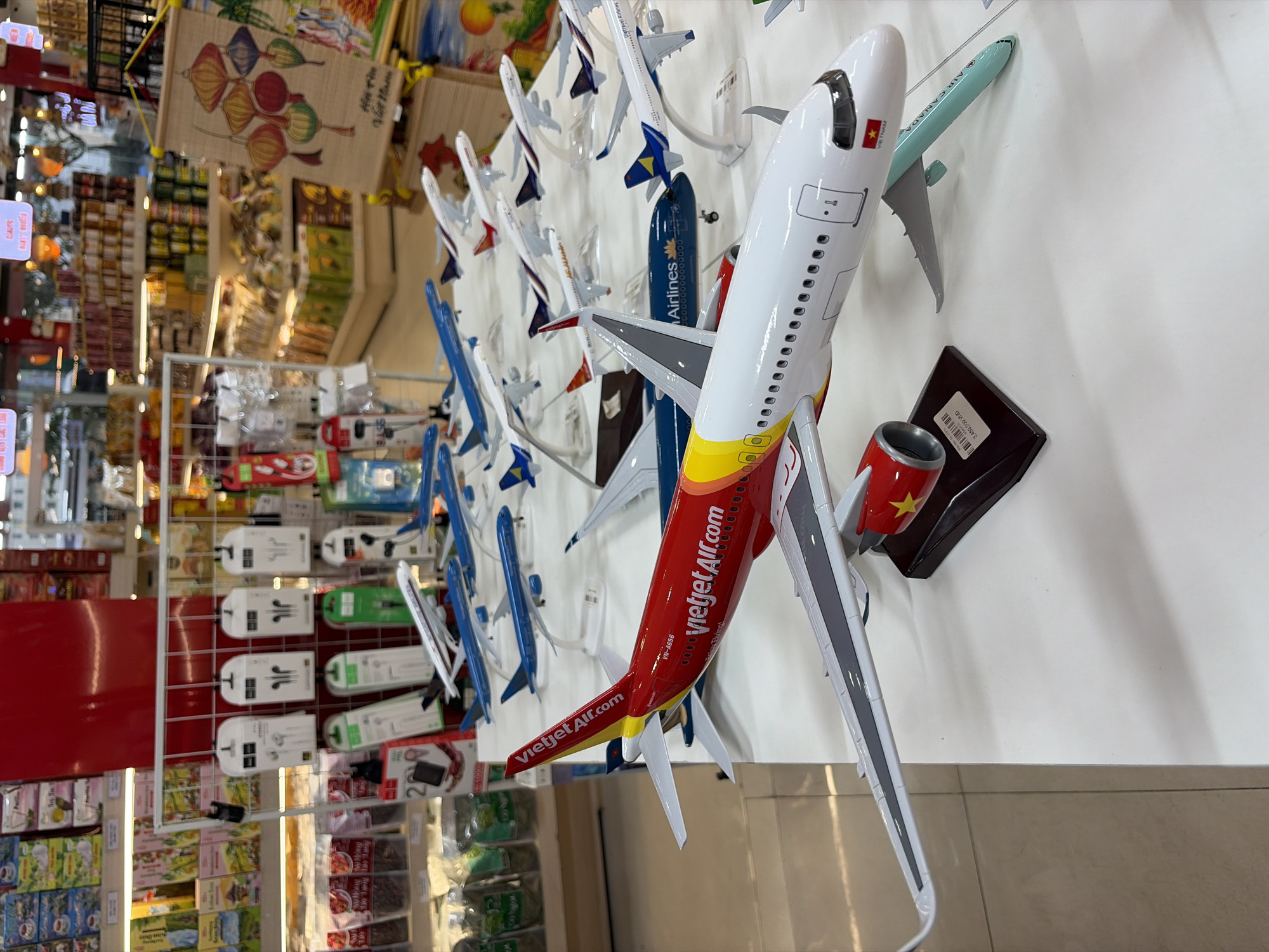The image shows a collection of airplane models on display, likely in a store or gift shop. The models are arranged on a white surface, and the background suggests an indoor environment with shelves of products, possibly in a market or retail setting in Đà Nẵng, Vietnam. The foreground features a prominent red and yellow Vietjet Air airplane model, with other airline models, including Vietnam Airlines, visible behind it. The scene is brightly lit, and the focus is on the detailed models of various aircraft.