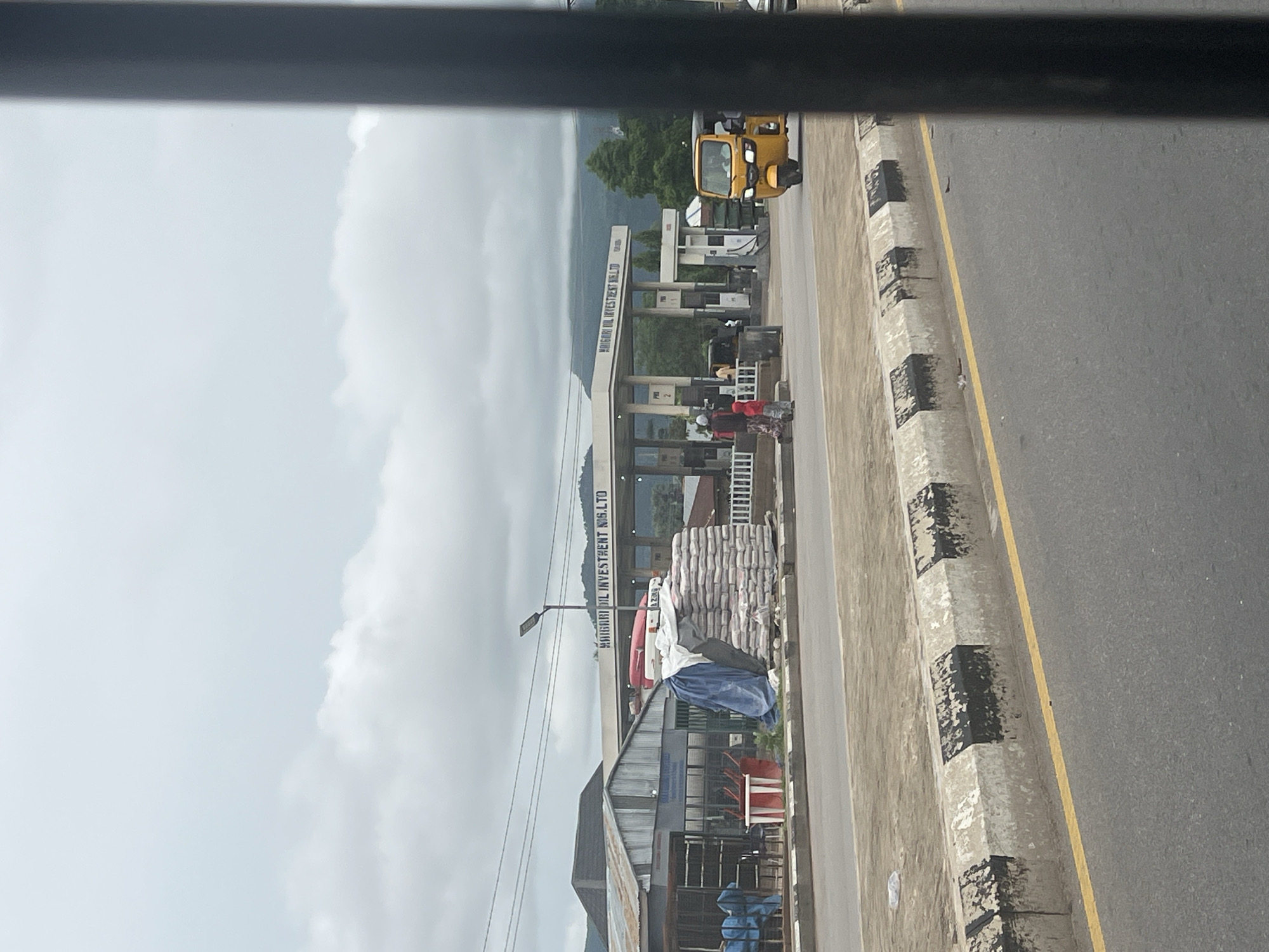 The image depicts a scene in Jalingo, Nigeria, featuring a roadside view of a gas station and surrounding structures. In the foreground, a paved road runs horizontally, with a yellow line marking its edge and a concrete curb painted with alternating black and white stripes. Beyond the curb, a strip of dry, dusty ground is visible.
Dominating the midground is a gas station with a white canopy supported by pillars. The name "MAIGORI OIL INVESTMENT NIG. LTD" is displayed on the canopy. Several fuel pumps are visible under the canopy. To the left of the gas station, a building with a corrugated metal roof and grey siding is present. Piles of bagged goods, likely cement or similar material, are stacked and partially covered by tarpaulins in front of this building. Red and white plastic chairs are also visible near the building.
Several people are present in the scene. Two individuals, one wearing a red jacket and the other in a patterned dress and headscarf, are walking near the gas station. Another person appears to be operating a yellow three-wheeled vehicle, commonly known as a "Keke" or "Marwa," which is driving on the road. Other individuals are sparsely visible near the gas station pumps.
The background reveals a slightly overcast sky with diffused clouds. Lush green trees and hilly terrain can be seen in the distance, suggesting a location with natural landscapes surrounding the urban environment. The overall atmosphere appears to be a typical roadside scene in a Nigerian town or city.