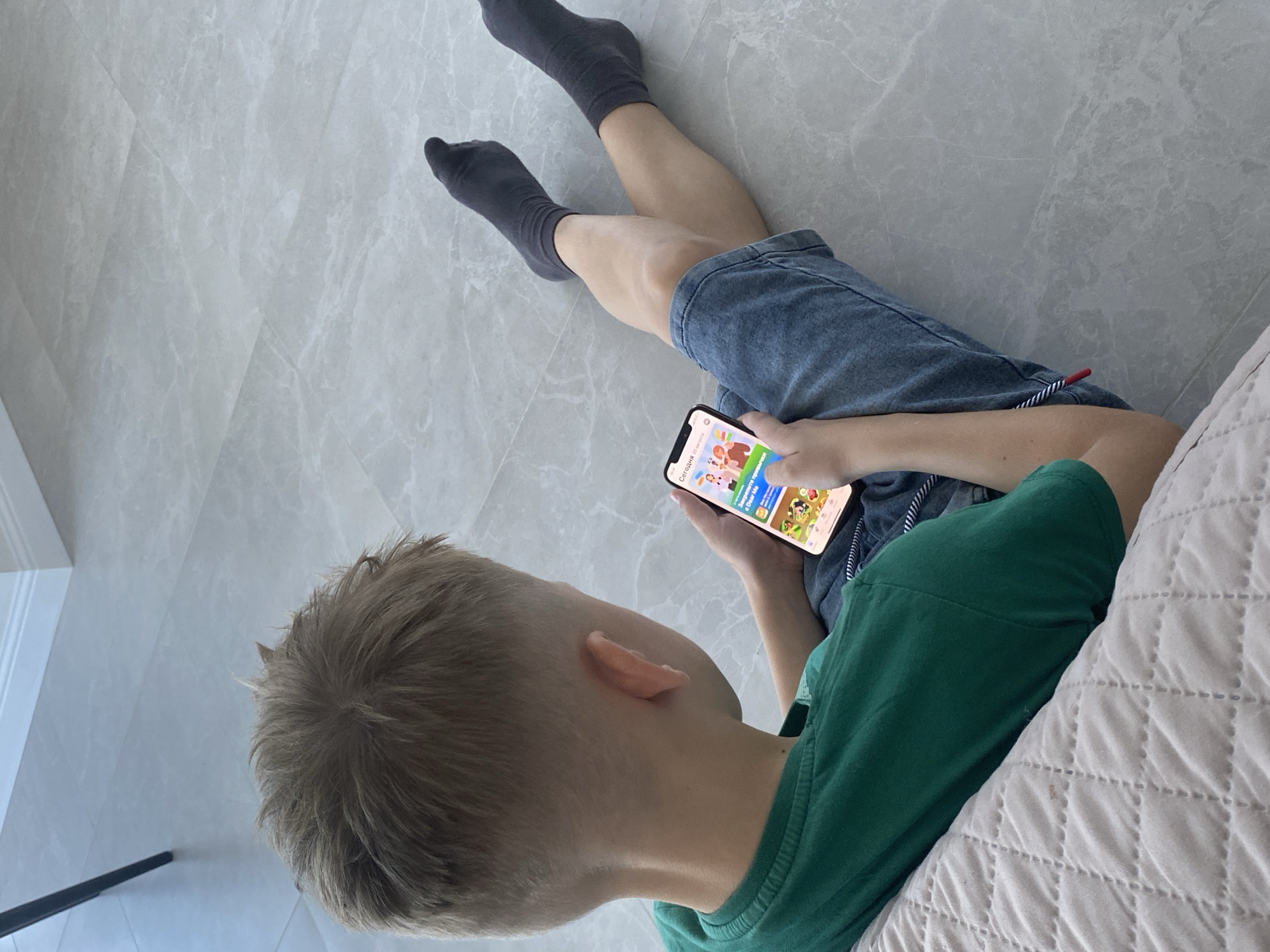 The image shows a person, likely a child, lying on a tiled floor, playing a game on a smartphone. The individual is wearing dark gray socks and denim shorts. Their torso is covered by a green t-shirt. The setting appears to be indoors, with a light-colored, patterned tile floor. A textured, light-colored surface, possibly a bed or couch, is visible in the lower right portion of the image.

The smartphone screen displays a colorful game interface with cartoonish characters. Visible text on the screen includes "Сегодня" (Today) and "в Идеи М" (in Ideas M), suggesting a Russian language interface. The time of day cannot be determined.

The lighting in the image is bright and even, indicating it might be daytime. The overall mood is relaxed and focused, as the person is engrossed in the game. The background is minimally detailed, focusing the viewer's attention on the subject and the activity. The person's hair is light brown and appears to be short. The angle of the shot is from above and to the side of the person, giving a clear view of their activity.