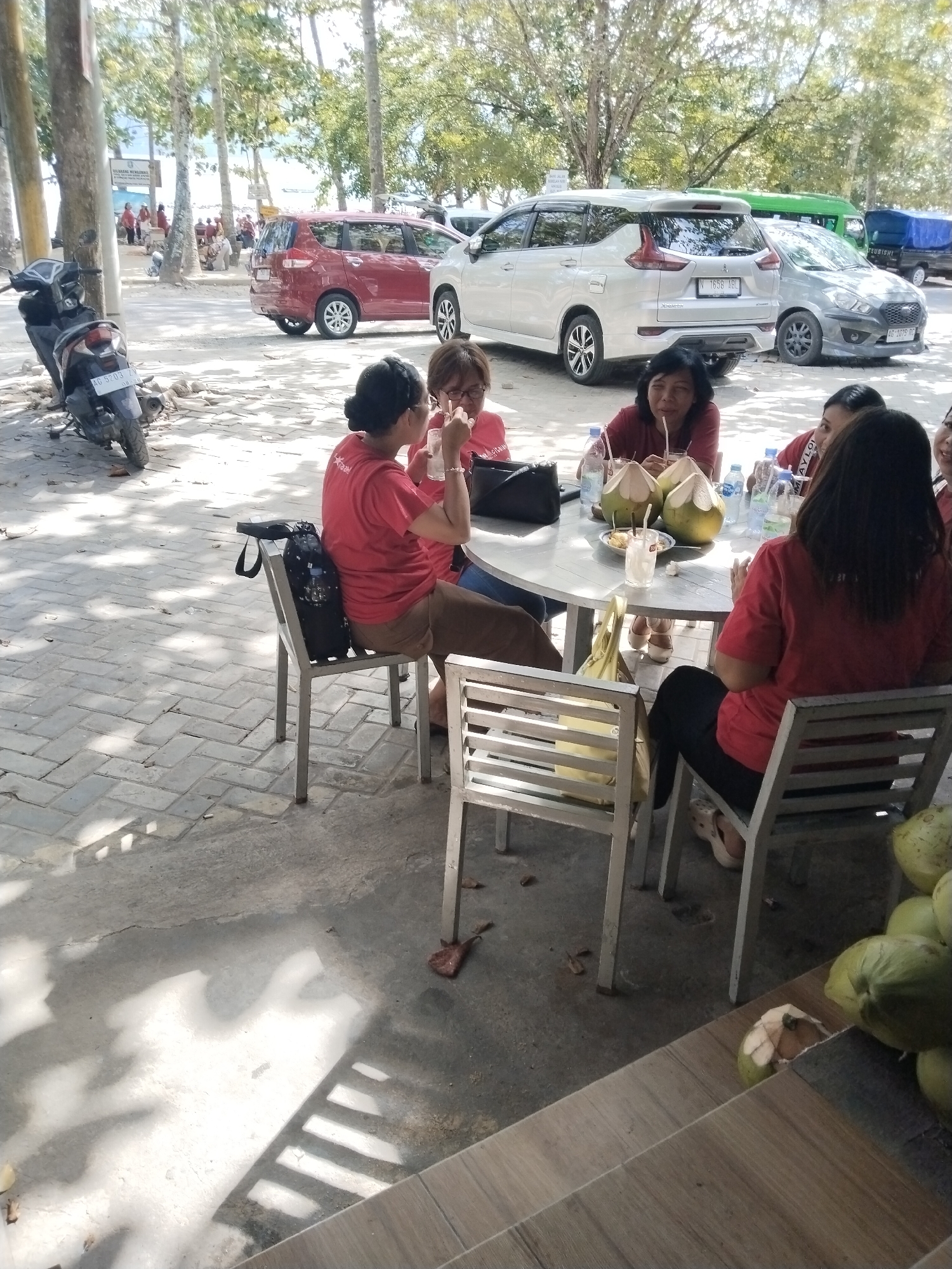 The image shows a group of people sitting around a table at what appears to be an outdoor cafe or restaurant in Prigi, Indonesia, near the beach. Several individuals are wearing red t-shirts, suggesting they might be part of a tour group or staff. They are consuming drinks, some of which are in clear plastic cups with straws. Two whole coconuts and several cut coconuts are present on the table, indicating they are likely drinking coconut water. A scooter is parked to the left of the scene, and several cars are visible in the background, including a red minivan, a silver SUV, and a gray hatchback. The environment appears to be warm and sunny, with palm trees and other foliage in the background, consistent with a coastal location in Indonesia. The ground is paved with interlocking bricks, and shadows from trees are cast across the area.