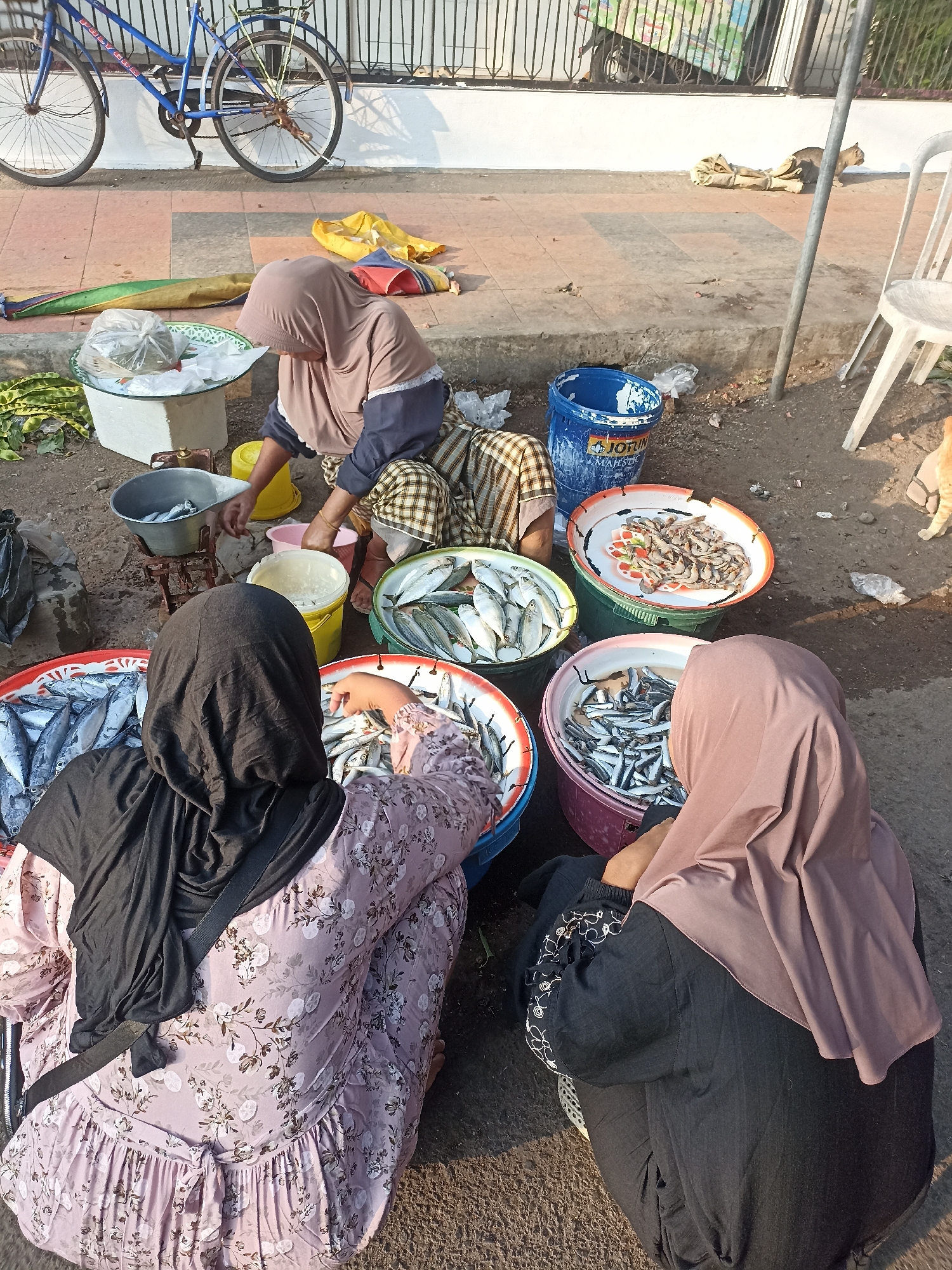 In Kelurahan Kepatihan, Indonesia, an outdoor market scene unfolds with three women engaged in selling fish. The women are dressed in hijabs and modest clothing. Two women are seated on the ground, sorting and displaying fish in large circular trays. The trays are filled with various types of small, silver-colored fish and some shrimp. A third woman is standing behind them, actively working with the fish. Various containers, including buckets and basins, are present, along with bags and what appear to be weighing scales. In the background, a blue bicycle is parked, and a cat is visible on a sack. The ground is paved with concrete and dirt, with some debris scattered around. The lighting suggests it is daytime, possibly morning or late afternoon, indicated by the long shadows. The overall impression is a busy, functional marketplace.