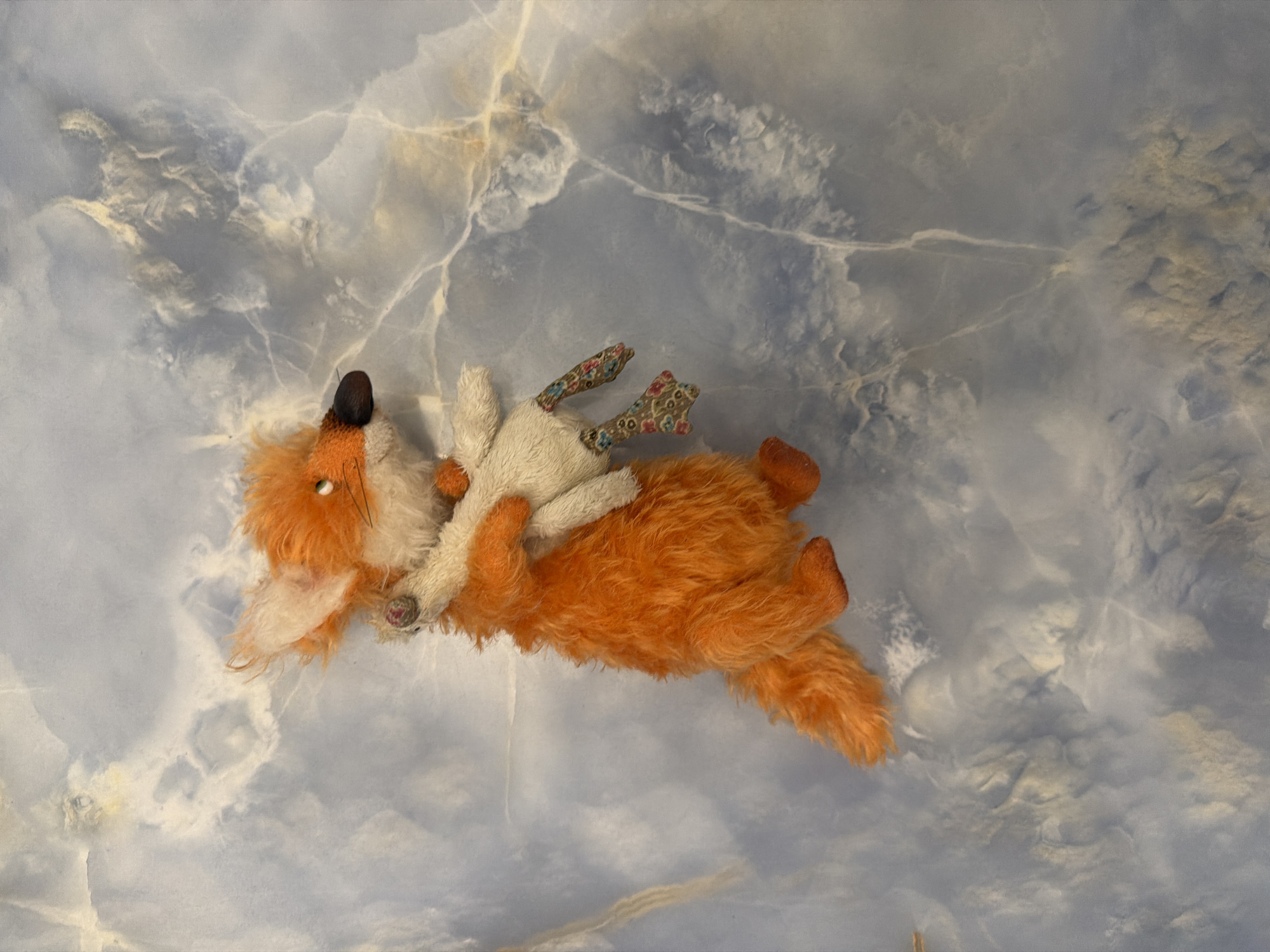 A plush toy fox is held from behind by a white plush toy, against a background of swirled blue and white marble. The fox toy is orange with a fluffy tail and ears, a black nose, and beady eyes. Its paws are visible, one of which is holding onto the white toy. The white toy is smaller and has floral-patterned legs. The marble background has white veins and appears to be a polished surface.