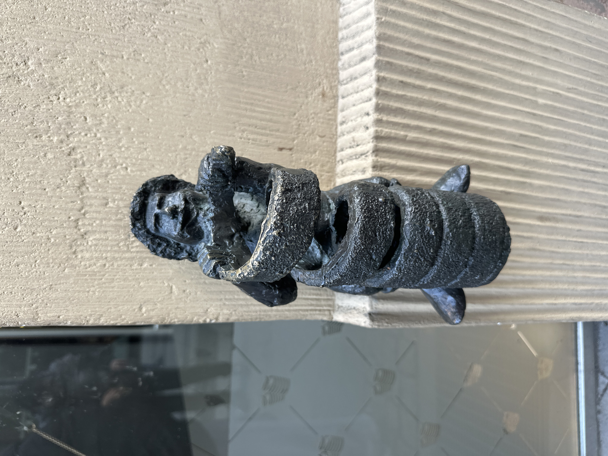 A close-up shows a metal door knocker affixed to a wooden door. The knocker is a stylized black sculpture depicting a human figure bound by coils, possibly representing a prisoner or captive. The door is light brown, with a textured surface on the right side. Below, there's a glass panel with a repeating geometric pattern. Based on its distinctive style and location, it is likely a detail from one of the ornate doors or gates found in the city of Wrocław, Poland.