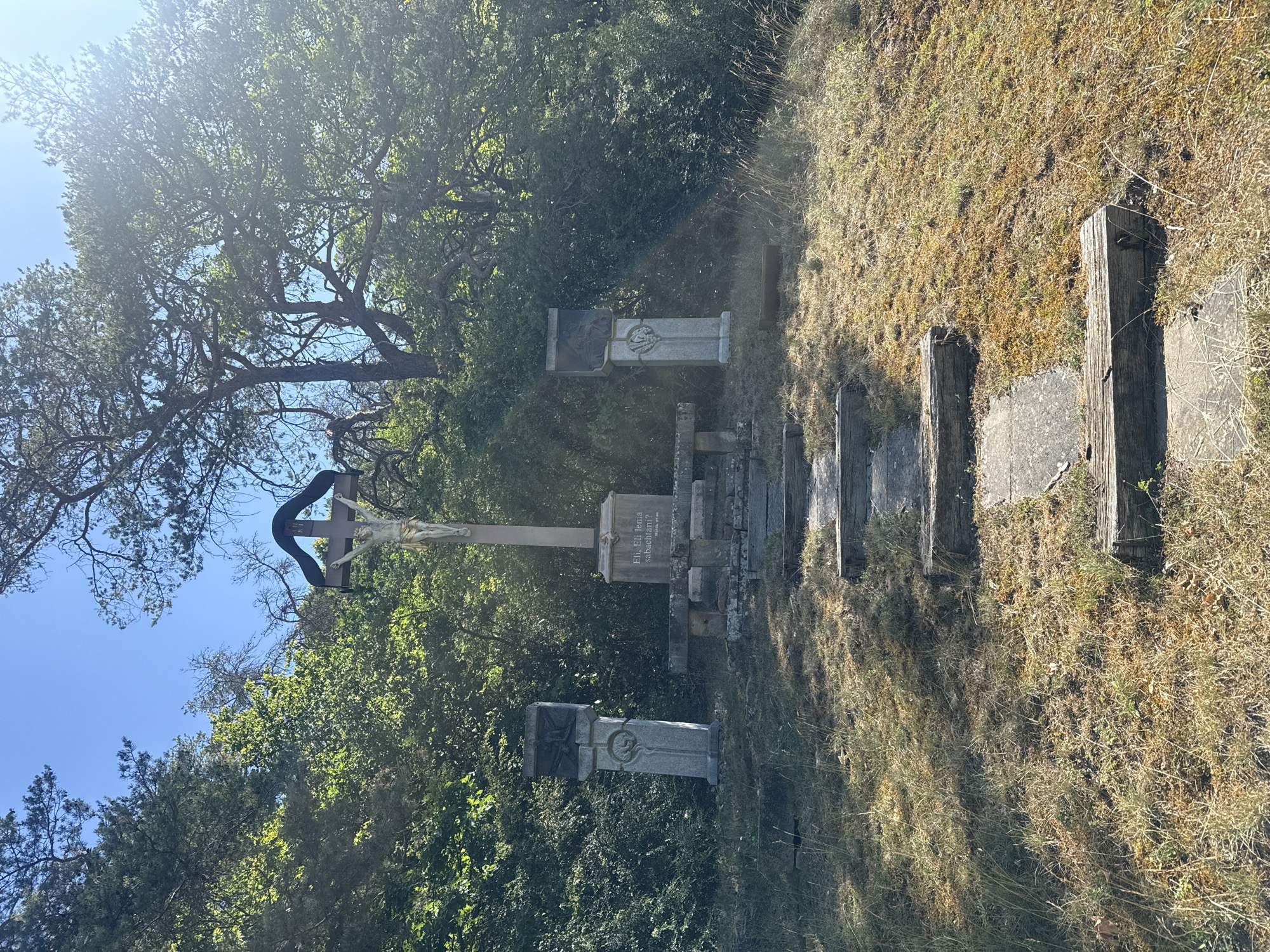 The image shows a religious monument located in Bad Salzschlirf, Germany. The monument consists of a large wooden cross with a figure of Jesus Christ, a stone base with inscriptions, and two smaller stone pillars on either side. The monument is situated on a grassy hill surrounded by trees. There are several steps leading up to the monument. The sky is clear and blue, and the sun is shining. The scene appears to be a place of worship or remembrance.