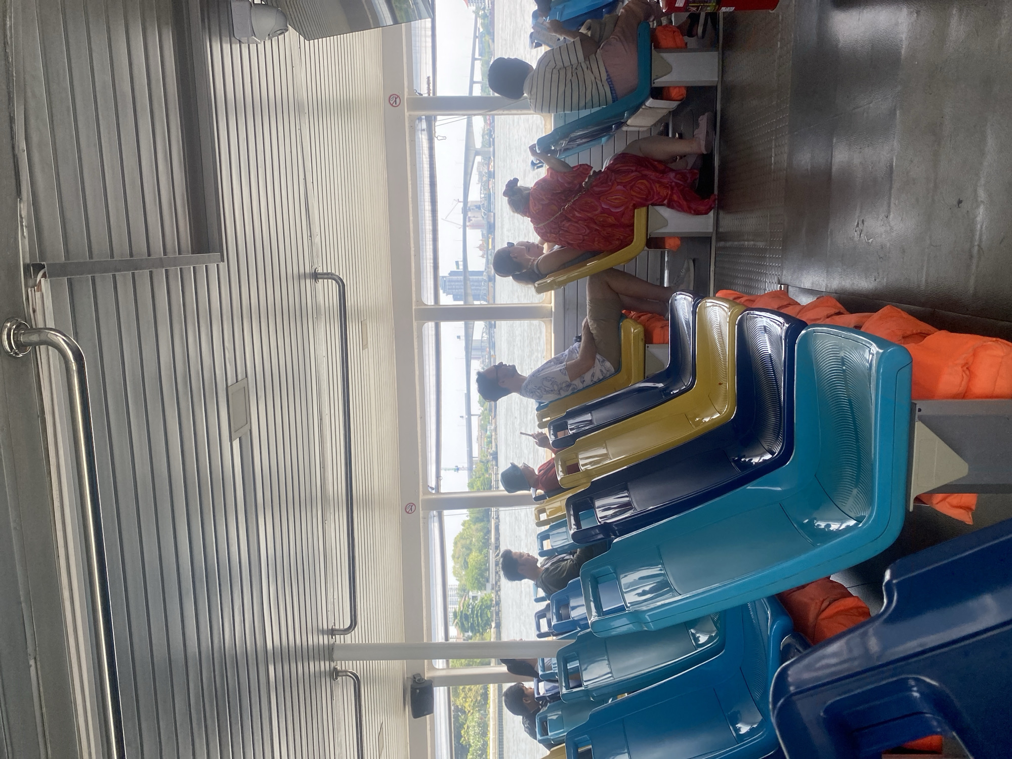 The image depicts the interior of a passenger ferry traveling on a river in Bangkok, Thailand. Passengers are seated in rows of blue and yellow plastic seats, with orange life vests visible beneath some seats. A woman wearing a red dress is sitting, and other people are seen sitting and looking out the windows. In the background, a bridge and buildings can be seen through the windows, indicating the ferry is on a river within an urban area. The ferry's interior has a metal wall with vertical panels.