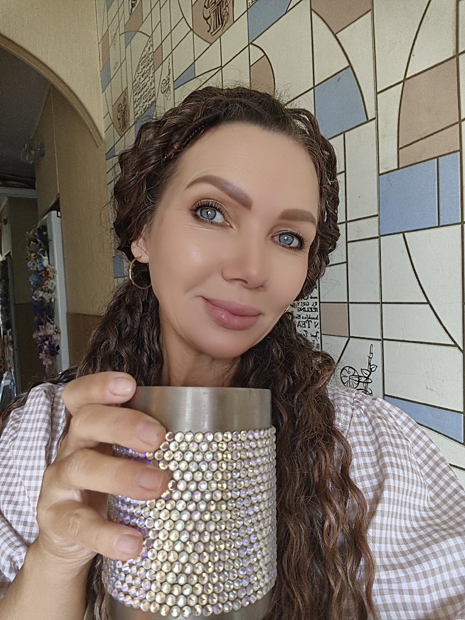 A medium shot shows a woman with long, curly brown hair holding a silver mug adorned with rhinestones. She is wearing a checkered shirt. The background features a wall with a mosaic-like pattern of geometric shapes and colors. The scene appears to be indoors, possibly in a home or cafe, with a hint of an arched doorway and decorations on the left side of the frame.