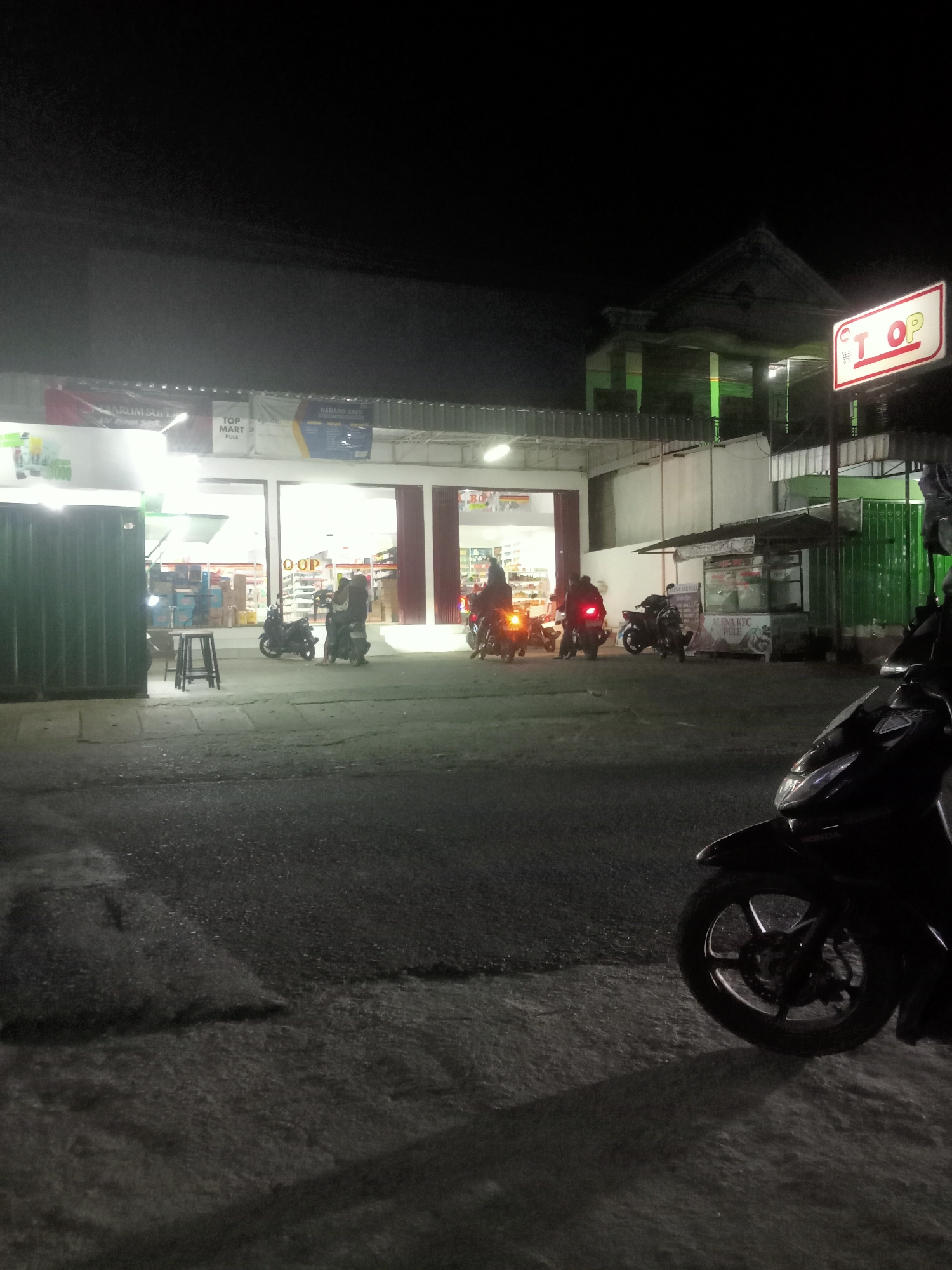 A nighttime street scene in Pule, Indonesia, captures the exterior of a brightly lit "TOP MART Pule" convenience store. The store's facade features large glass windows revealing shelves stocked with products, and a prominent red and white "TOP" sign is visible on the right. Several motorcycles are parked or in motion on the asphalt road in front of the store, with some riders visible. A small food stall with a green awning is situated to the right of the mart. The environment is dark, with artificial lighting from the store and streetlights illuminating the area.