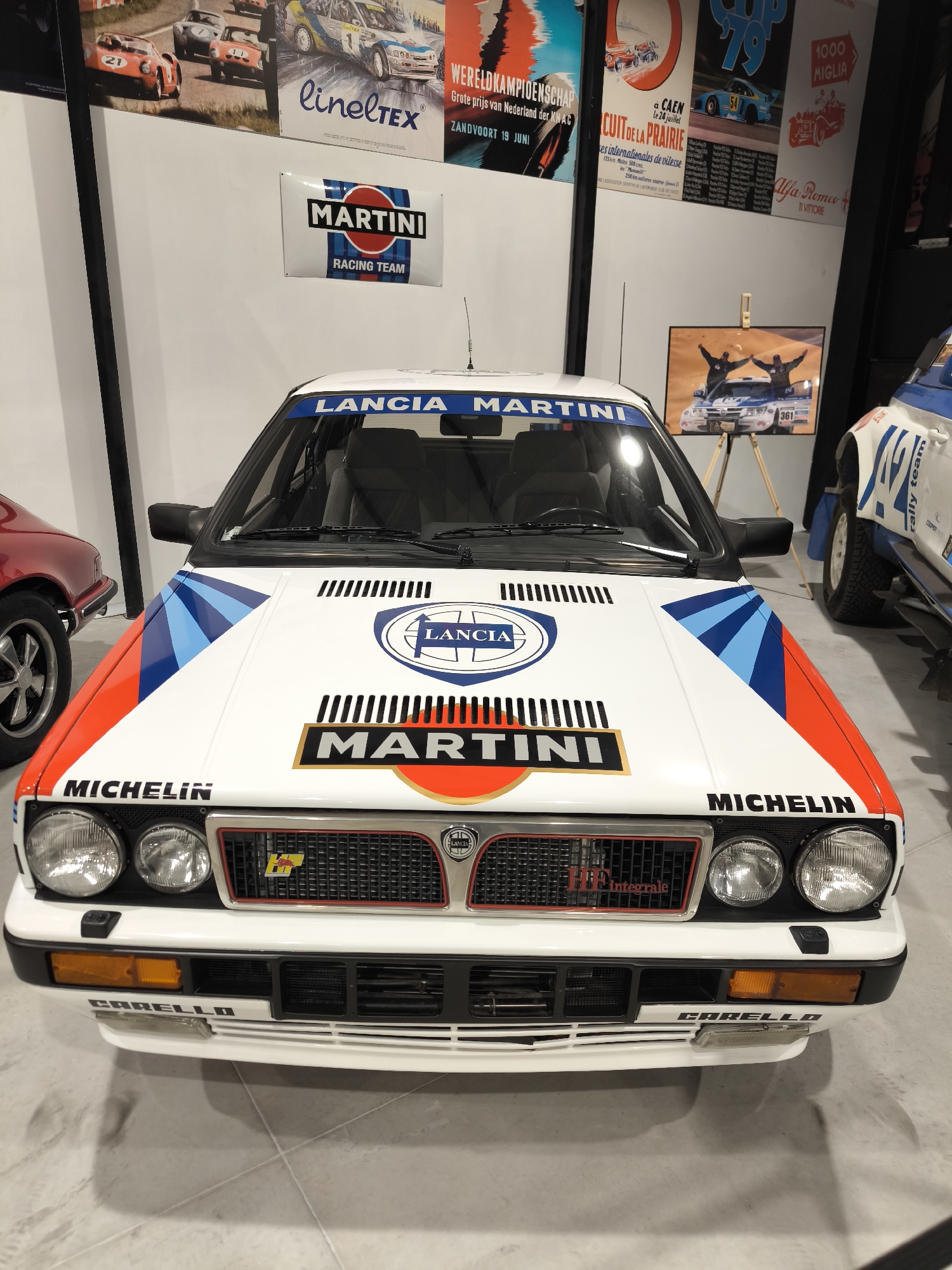 The image is a close-up shot of a white Lancia Delta Integrale rally car with Martini Racing livery. The car is indoors, likely in a museum or car collection, as evidenced by the clean, well-lit environment and the presence of posters and another car in the background. The focus is on the front of the car, showcasing the Lancia logo, Martini stripes, and various sponsor decals, including Michelin and Carello. Other objects in the surrounding context include racing posters and a photograph on an easel. The photo was not taken in Kapatovo, Bulgaria.