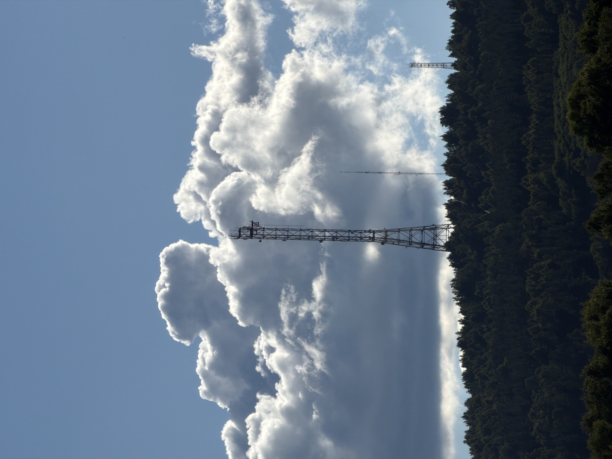 The image depicts a dramatic sky filled with large, billowing white and gray clouds. A tall, lattice tower, likely a radio or transmission tower, stretches across the frame, silhouetted against the bright sky. The tower is a prominent object in the scene. To the right, the dark green, densely packed foliage of a hillside or mountain range fills the lower portion of the image. The trees create a strong contrast with the light sky and clouds. There are also hints of other, more distant lattice towers visible in the sky, suggesting a landscape with telecommunications infrastructure. The lighting suggests it is daytime, likely mid-morning or afternoon, given the brightness of the sky and the way the clouds are illuminated. The clouds themselves appear dynamic, with some areas intensely white and others a darker gray, hinting at possible weather activity or varying atmospheric conditions. The scene is naturalistic and seems to capture a moment of striking cloud formations juxtaposed with man-made structures and a natural landscape. No text is visible in the image.