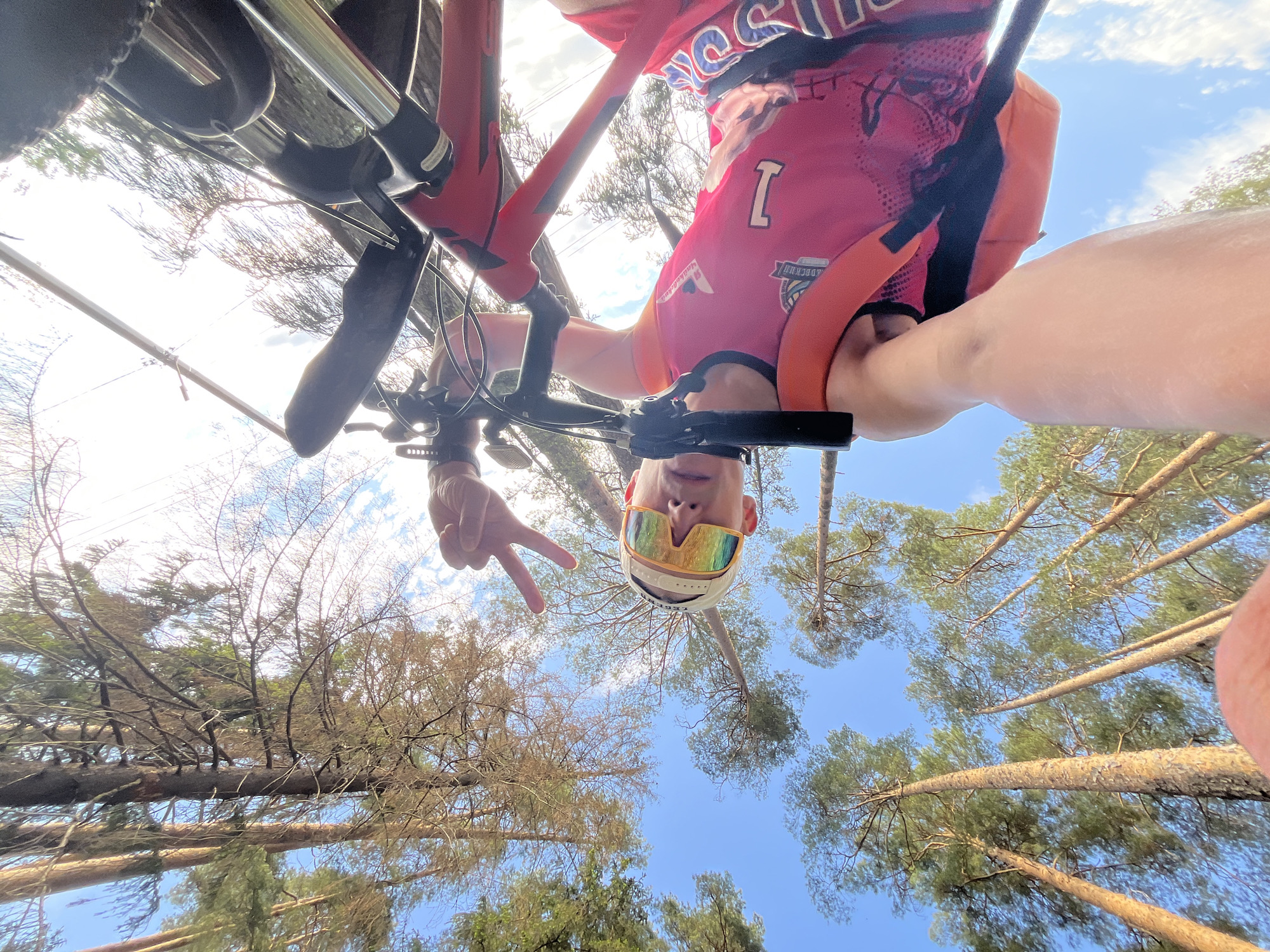 A person wearing a cycling jersey, helmet, and sunglasses is holding a bicycle and making a peace sign. The photo is taken from a low angle, looking up into a forest with a blue sky visible between the trees. Based on the jersey design, it is plausible this image was taken in or around the city of Chaikovsky, Russia.