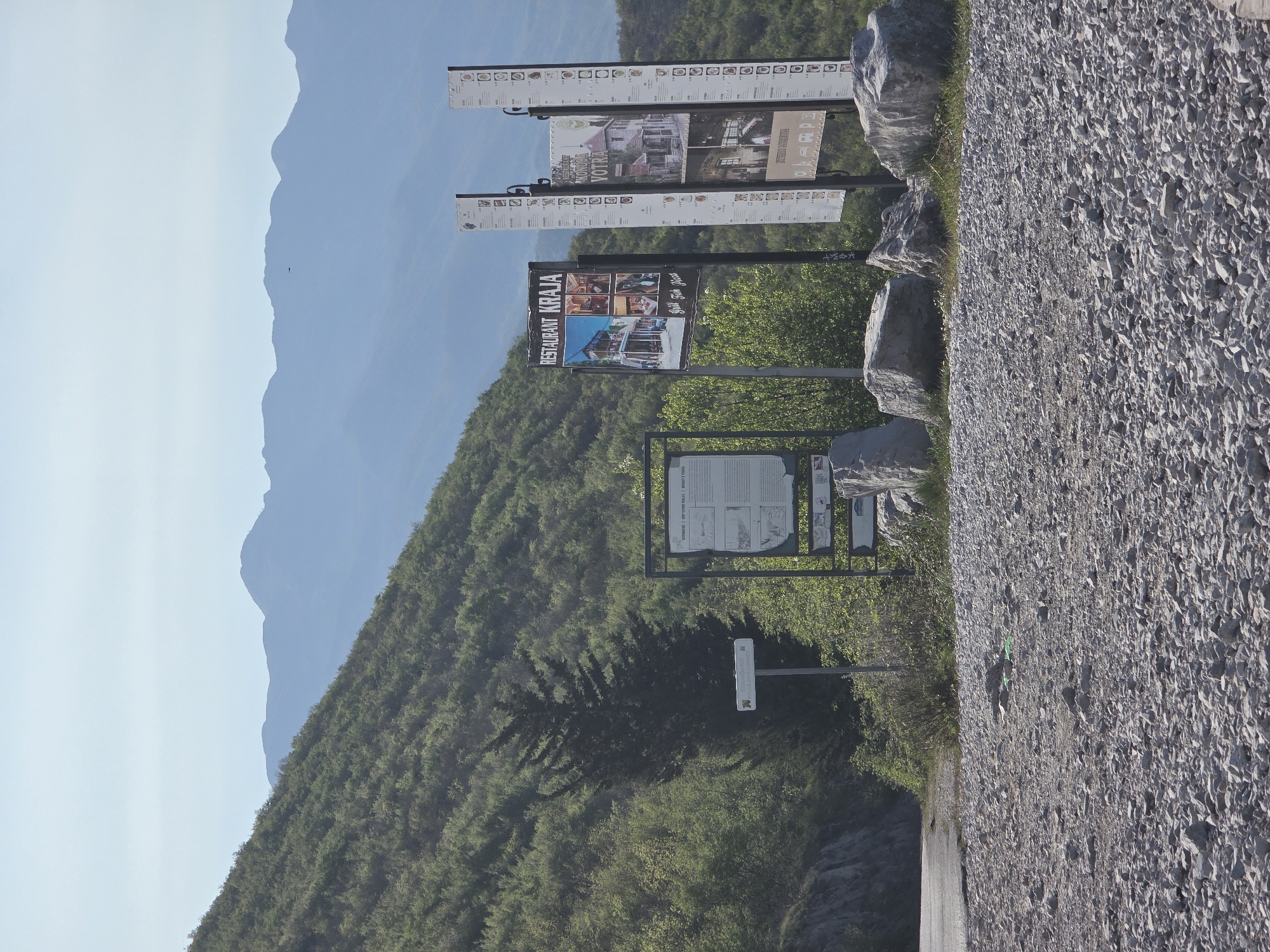 The image shows a roadside view in Arbnež, Montenegro. Several informational signs are visible, including a sign for "RESTAURANT KRAJA" with food images, and signs displaying historical and cultural information. The signs are arranged amongst several large rocks. In the background is a lush green hillside leading to a mountainous terrain. The roadside is edged by a gravel surface. The sky is pale blue with some wispy clouds.