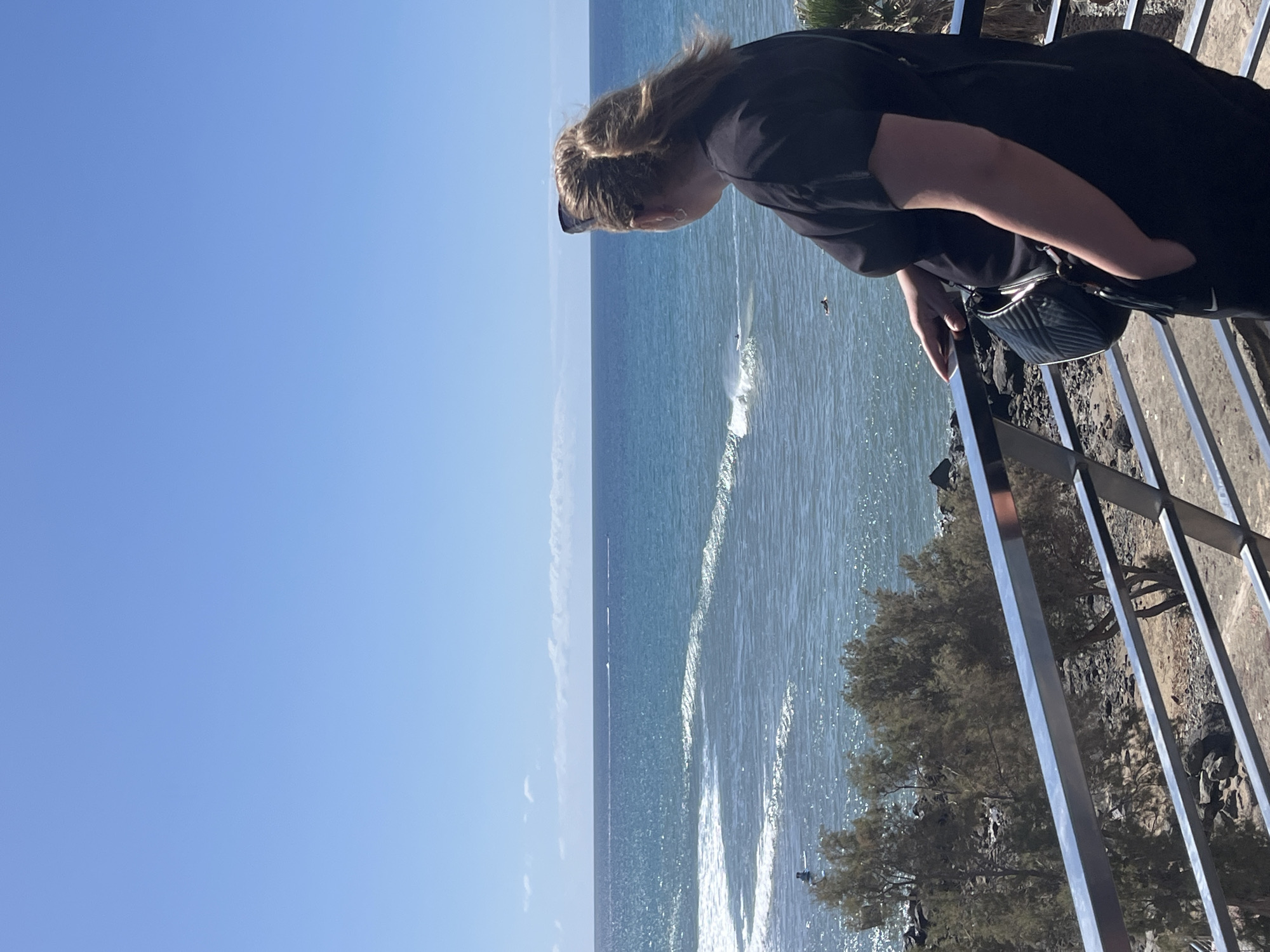 A woman in a black top leans over a railing, observing the ocean. The sky is clear blue. The ocean has visible waves. In the background, a faint landmass can be seen on the horizon. The railing is made of vertical metal bars. A tree is visible behind the railing. The location is Maspalomas, Spain.