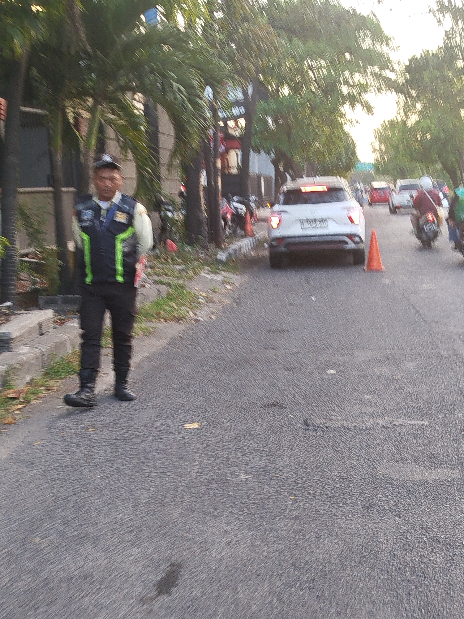 A person wearing a security uniform with reflective green and black accents, a light-colored long-sleeved shirt, dark pants, and black boots stands on an asphalt road. The individual is facing slightly to the left and appears to be observing the surroundings. Behind the person, several palm trees line the street, with buildings and other vegetation visible further in the background. Several vehicles, including a white SUV, motorcycles, and other cars, are present on the road, suggesting a street or roadway in a city environment. An orange traffic cone is placed on the road near the white SUV. The lighting suggests it is daytime, possibly late afternoon, with sunlight filtering through the trees. The overall scene depicts a street in Surabaya, Indonesia, where the security person is likely on duty.