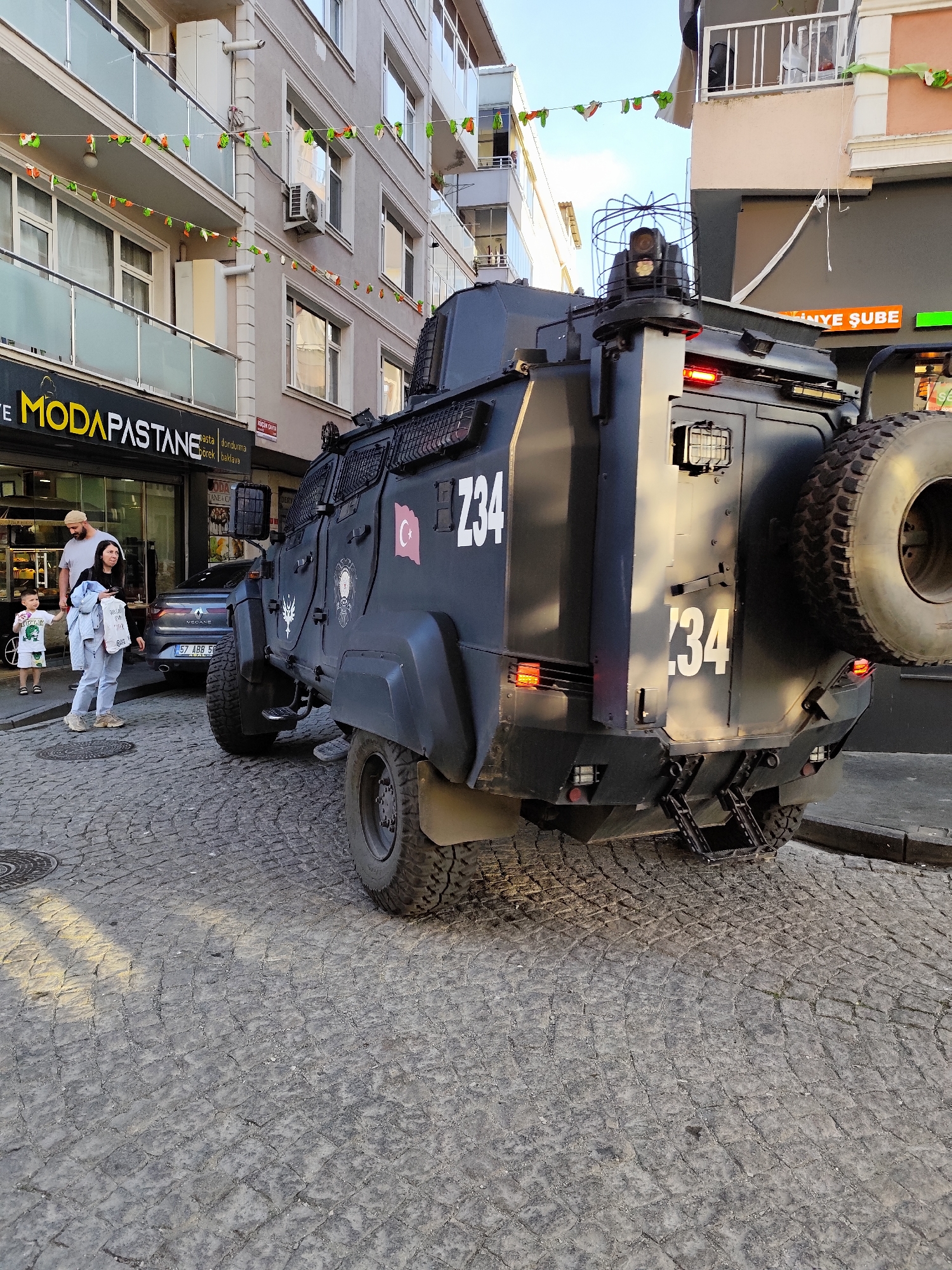 The image depicts the back and side of a dark armored military vehicle with the number "Z34" prominently displayed on its exterior, parked on a cobblestone street in Sarıyer, Turkey. A spare tire is mounted on the back. A Turkish flag is on the side of the vehicle.
In the background, a family of three—a man, a woman, and a child—stand near a storefront labeled "MODAPASTANE," which offers pastries, ice cream, and baklava. A gray Renault Megane is parked in the background next to the vehicle. The street is decorated with colorful pennants. The scene is set during daylight, casting shadows on the cobblestone street. The buildings around the storefront are multi-story structures with windows and air conditioning units.