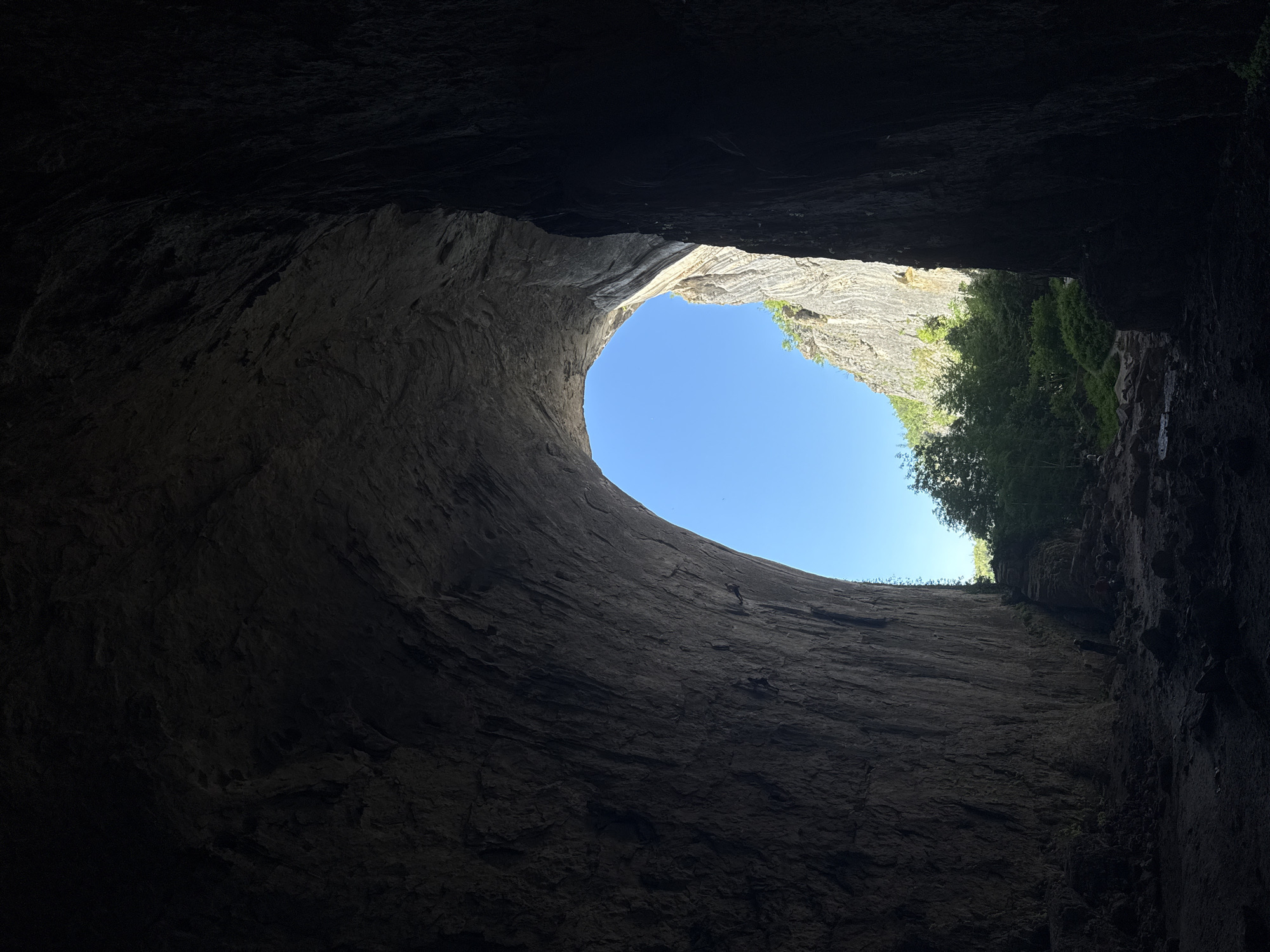 The image captures the view from inside the Prohodna Cave, also known as the "Eyes of God," in Karlukovo, Bulgaria. The perspective is from the dark interior of the cave, looking out towards a bright, open sky. The cave walls are massive and textured, with the opening forming a distinct, large archway.
Dominating the foreground is the rough, textured rock of the cave's interior, rendered in dark, shadowed tones. The scale of the rock formations is evident, showcasing their geological ruggedness. As the eye moves towards the center and right side of the frame, the darkness gives way to the immense arch of the cave's entrance.
Through this arch, a vibrant blue sky is visible. Beyond the immediate entrance, a lush green landscape unfolds, characterized by dense trees and foliage. The sunlight illuminates the surrounding cliffs, highlighting their layered rock structure. The contrast between the dark, enclosed space of the cave and the bright, open expanse outside is striking.
In the lower right section of the cave floor, near the transition to the outside, a few silhouetted figures are visible. They appear to be small in comparison to the cave's scale, suggesting they are visitors or individuals exploring the area. The ground inside the cave is uneven, composed of rocks, dirt, and debris, indicative of a natural, untamed environment. The overall scene evokes a sense of awe and the grandeur of nature.