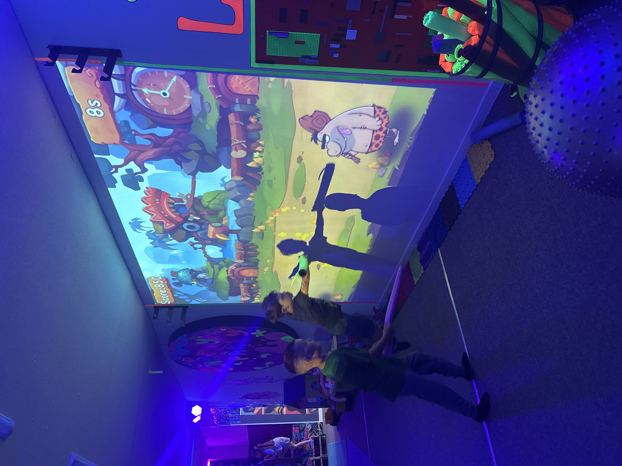 Here is a detailed description of the image:
**Scene:** The image captures an indoor interactive play area.
**People:** Two children are present, actively engaged in an interactive game projected onto a wall.
**Objects:**
* **Projected image:** A large, cartoon-style image is projected onto the wall. The image features a prehistoric setting with a caveman character, a clock, and game elements.
* **Foam Toys:** There are foam tubes and other soft play equipment on the right edge of the image.
* **Other decorations:** Various colorful decorations adorn the room, including patterns on the walls and flooring.
* **Ball:** A large, textured ball is located on the right side of the image.
**Actions:** The children are interacting with the projected image, possibly aiming a toy or using their movements to control the game on the screen. Their shadows are cast onto the screen.
**Environment:** The room appears to be dimly lit with a blue-purple hue, giving it a vibrant atmosphere. The space is clearly designed for children's play and interactive activities.
**Location:** The city is noted as Unknown, Unknown.