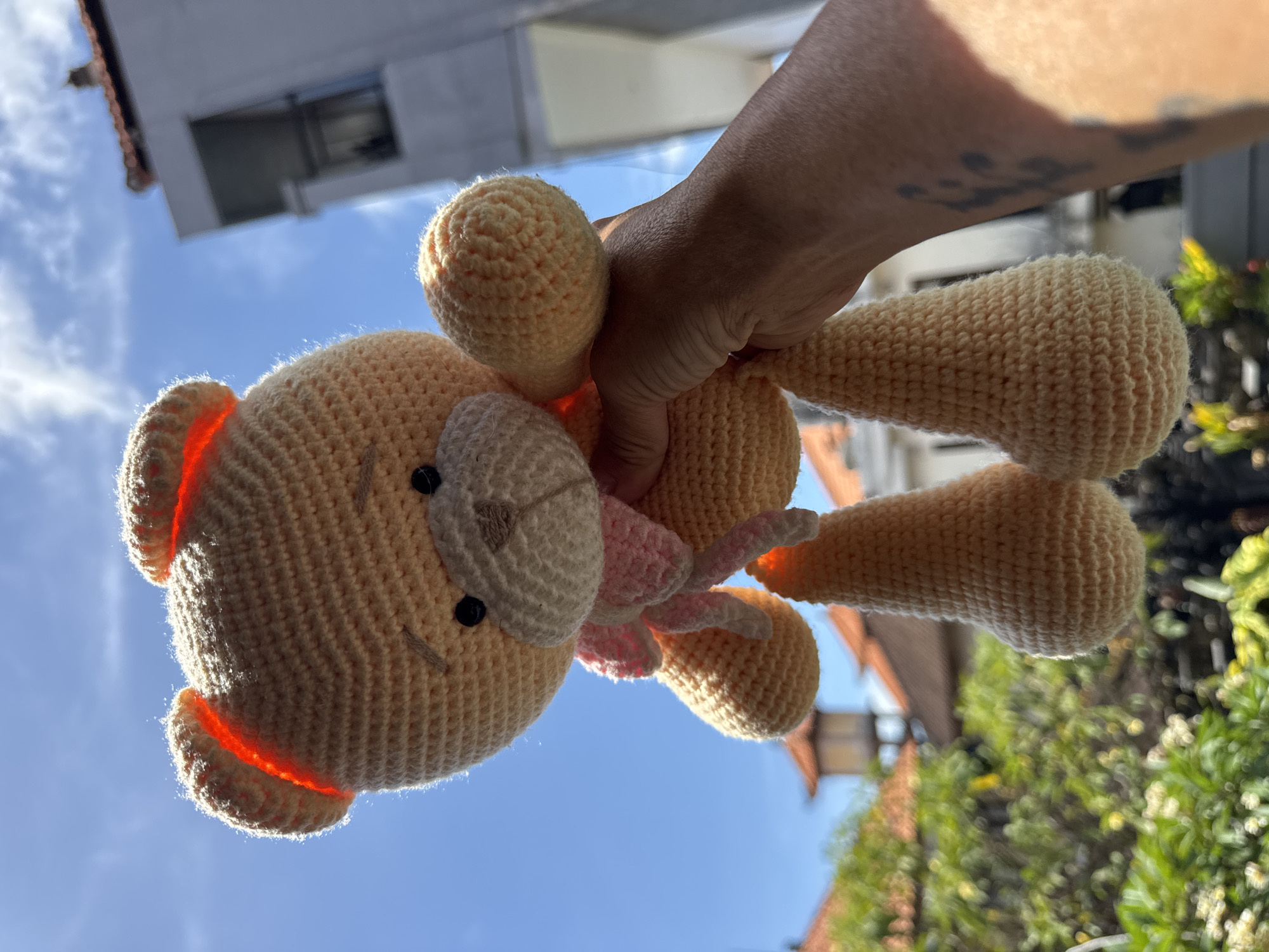 A person's hand is holding a crocheted, tan-colored stuffed animal, likely a bear, against a background of blue sky with some white clouds. The bear has a lighter tan face, dark eyes, and a pink bow. The background also includes parts of buildings and trees, suggesting an outdoor setting. The image was likely taken in Kuta, Indonesia, based on the presence of buildings and foliage typical of the area.