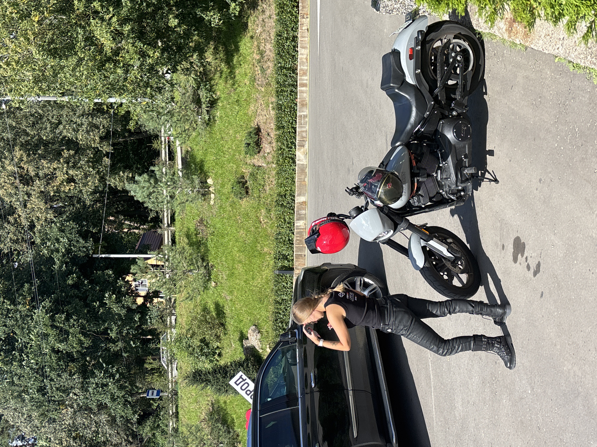 A blonde woman wearing a black tank top, black leather pants, and black combat boots stands next to a black SUV and a grey motorcycle. She has her right hand on her head, and her left hand is in her pocket. The motorcycle has two helmets on it, one red and one black with gold accents. The SUV has a white sign with red lettering that says "РОД" (Rod) attached to its roof rack. The scene takes place on a paved area next to a grassy embankment with trees. In the background, a white utility pole with power lines is visible. The setting is consistent with the outskirts of Zvenigorod, Russia, possibly near a park or roadside rest area.