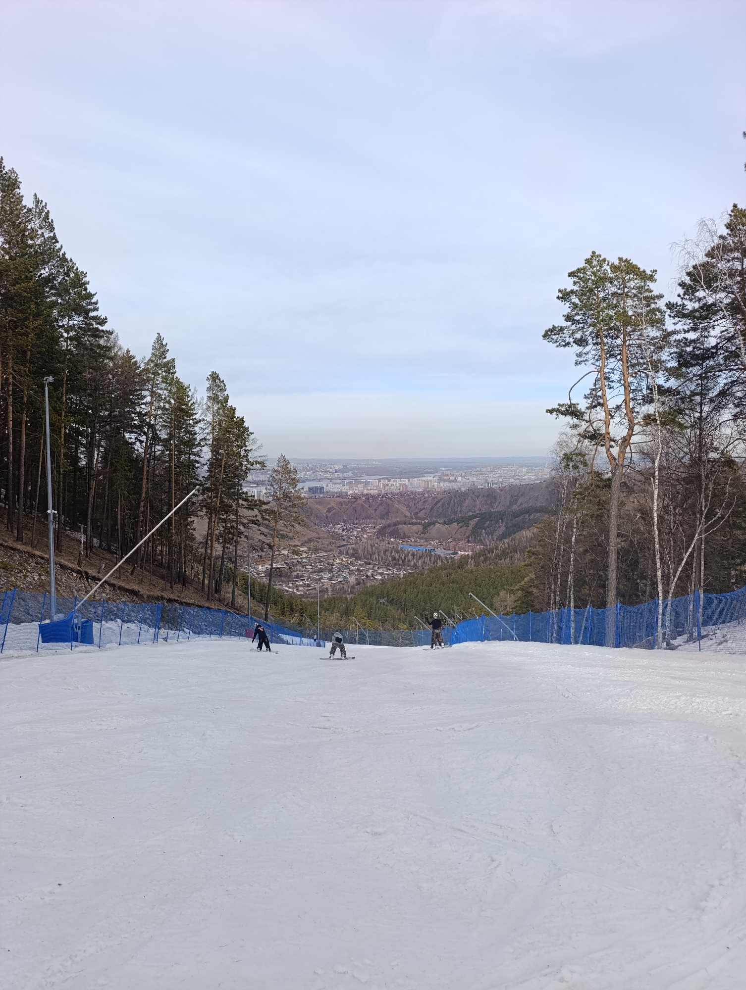 The image shows a snow-covered ski slope with a view of the city of Krasnoyarsk, Russia, in the distance. The scene is framed by coniferous trees on either side. A blue safety fence runs along the edges of the slope. Three people are snowboarding down the slope. Ski poles and a snowmaking machine are visible on the left side. The sky is overcast. The city of Krasnoyarsk is visible in the background, nestled in a valley.