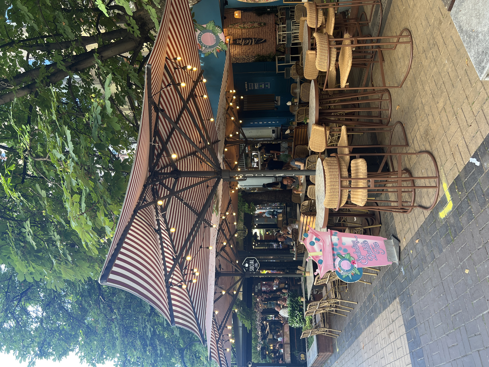 This media file depicts an outdoor seating area of a restaurant or cafe with a striped awning and string lights. The scene is bustling with people, suggesting it is open for business. The tables and chairs are made of wicker and metal, arranged on a brick-paved sidewalk. Lush green trees overhang the seating area, providing shade.
In the background, the establishment's interior is visible, showing more seating and a brick wall with decorative elements, including surfboards and a sign that reads "Aloha". A pink sign shaped like a bubble tea cup, advertising "Exploding Bubble Tea," is prominently placed on the sidewalk. Another sign reads "SOCIAL" with text below it.
The time of day appears to be daytime, likely late afternoon or early evening, given the lighting from the string lights and the shadows cast. The weather seems pleasant, suitable for outdoor dining. The overall atmosphere appears lively and social. The location context is Sofia, Bulgaria.