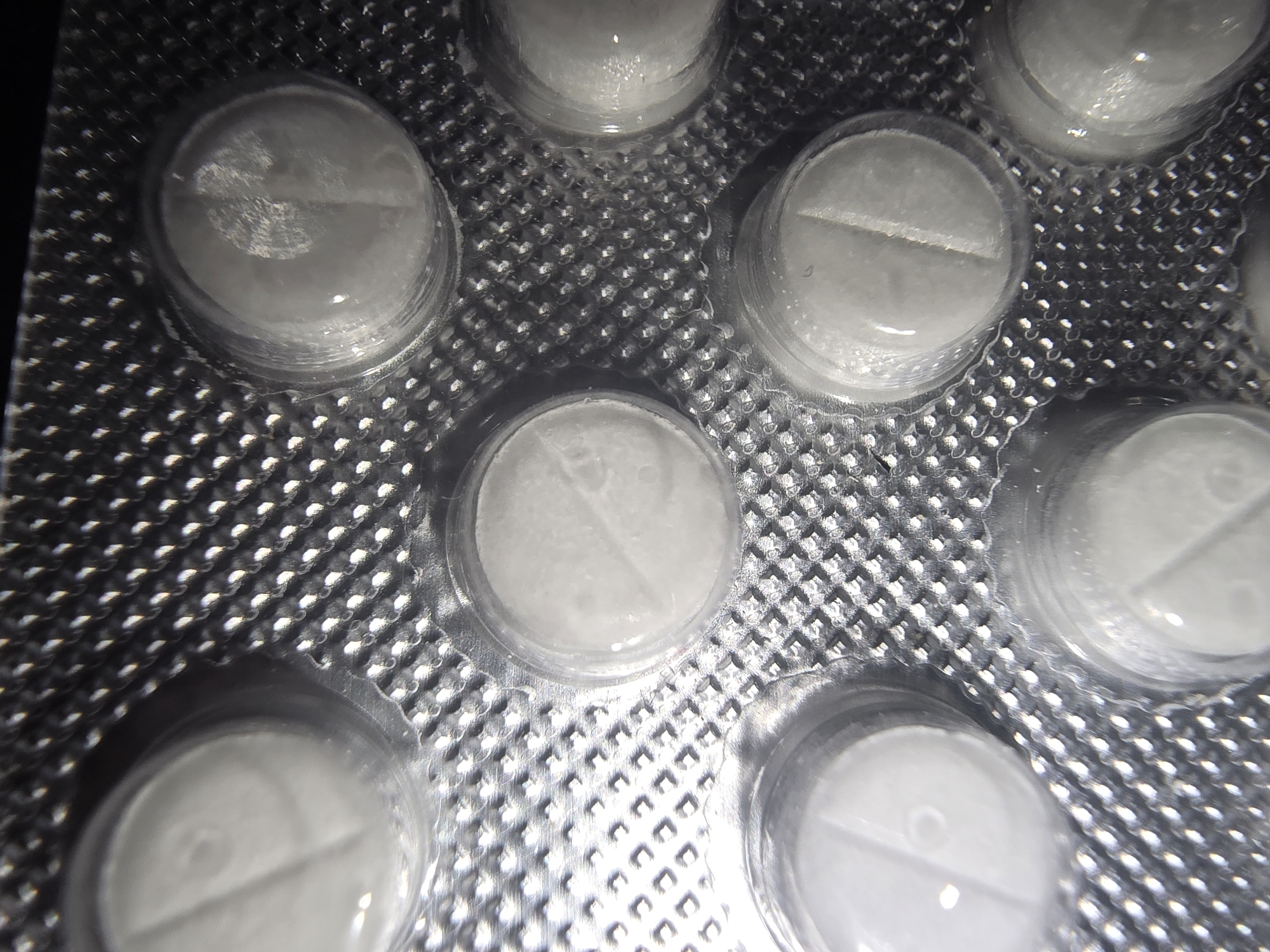 The image shows a close-up, top-down view of white pills in a silver blister pack. The blister pack is made of plastic and foil, with a textured pattern underneath the individual pill compartments. The pills are round and appear to have a score line down the middle, indicating they can be broken in half. The lighting is harsh, creating reflections and shadows on the pills and the packaging. The pills are arranged in a grid pattern within the blister pack. The image does not provide any information about the location of the pills in Ulan-Ude, Russia, or any details about people, actions, or interactions.