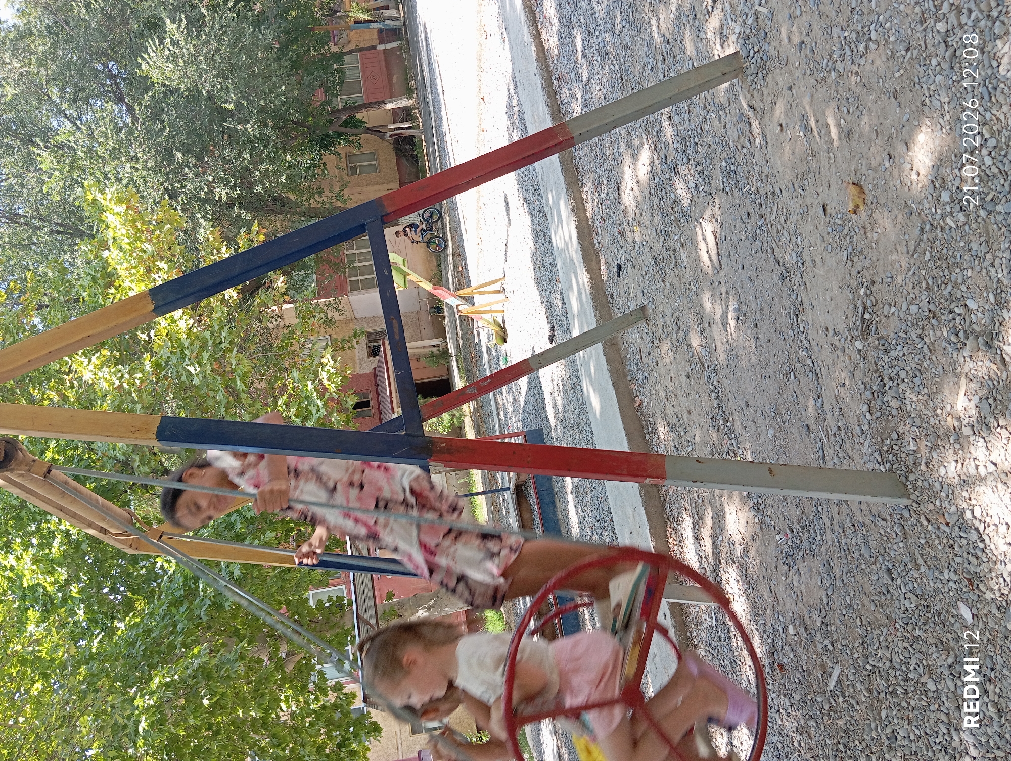 In Bekabad, Uzbekistan, two young girls are playing on a swing set in a park. The older girl, wearing a floral dress, is pushing the younger girl, who is in a pink skirt and white top, on a circular swing. The playground is covered with small stones and gravel. In the background, there are trees and apartment buildings. A young boy on a bicycle is visible further in the background. The date stamp on the image reads "21.07.2026 12:08".