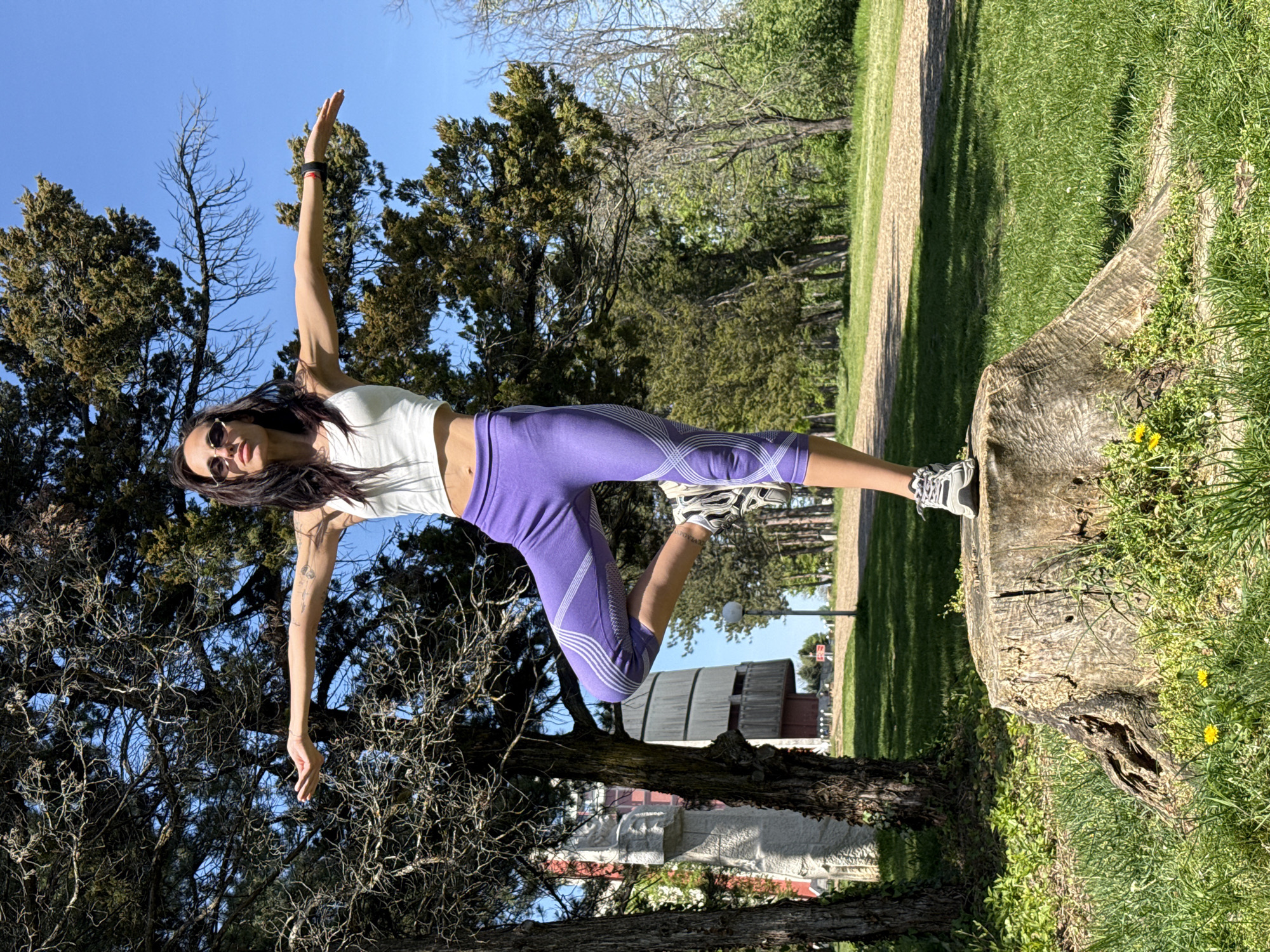 A young woman is captured in a dynamic balancing pose on a tree stump in a sunny outdoor setting, likely a park in Pleven, Bulgaria. She is performing a yoga or fitness-inspired movement, standing on one leg with the other bent behind her and her arms outstretched horizontally for stability.
The woman has dark hair and is wearing a white sleeveless crop top, purple capri-length athletic leggings adorned with white geometric patterns, and grey and white athletic shoes. She is also wearing sunglasses. Small tattoos are visible on her arms, and she has a black wristband on her right arm.
The scene is set under a clear, bright blue sky, indicating a sunny day, likely midday or afternoon given the strong shadows cast on the grass. The foreground features the rough, textured tree stump she is balancing on, surrounded by lush green grass. Various trees, including dense evergreens and bare-branched deciduous trees, populate the background, creating a natural and verdant environment. A dirt path winds through the park, and in the distance, a large, light-colored building, possibly an old barn or industrial structure, is visible amidst the foliage. The overall atmosphere is active and serene, emphasizing outdoor fitness. There is no visible text in the image.