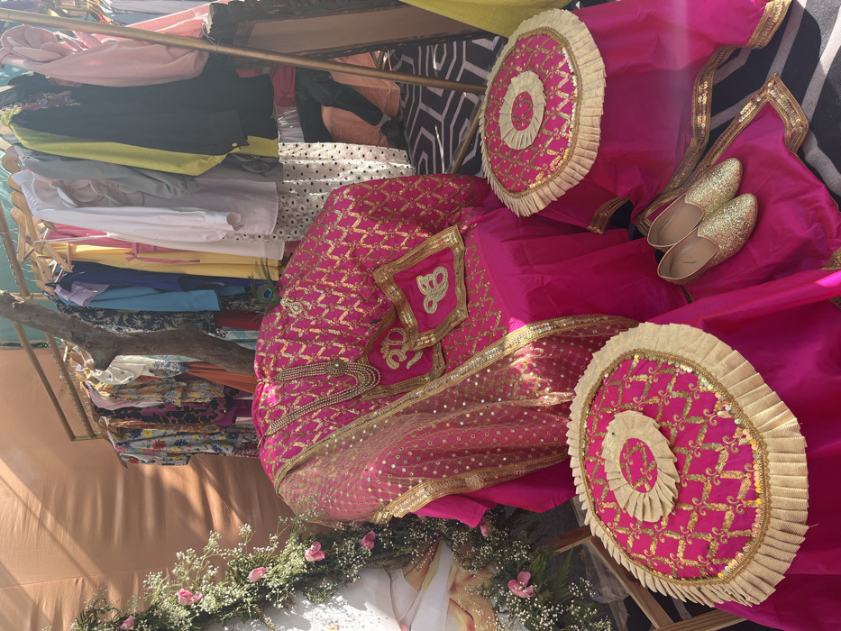 The image shows a close-up of a decorative wedding setup in a vibrant, outdoor setting. The scene is located in Sector 17, Chandigarh, India, at coordinates 30.764 latitude and 76.797 longitude. The centerpiece is a pink, sequined chair draped with a matching pink shawl adorned with gold embroidery. Two pink, sequin-covered cushions with gold embroidered accents are placed on the chair, and a small, ornate necklace rests on the seat. Two additional pink cushions with gold embroidery and ruffles are situated on the floor. Two pairs of golden shoes are placed on the pink fabric to the right of the chair. The chair and cushions are arranged on a black and white patterned floor. A rack filled with colorful clothing is visible in the background. The surrounding environment includes lush green foliage and a peach-colored wall. The image captures the elaborate decorative details often used in Indian weddings.