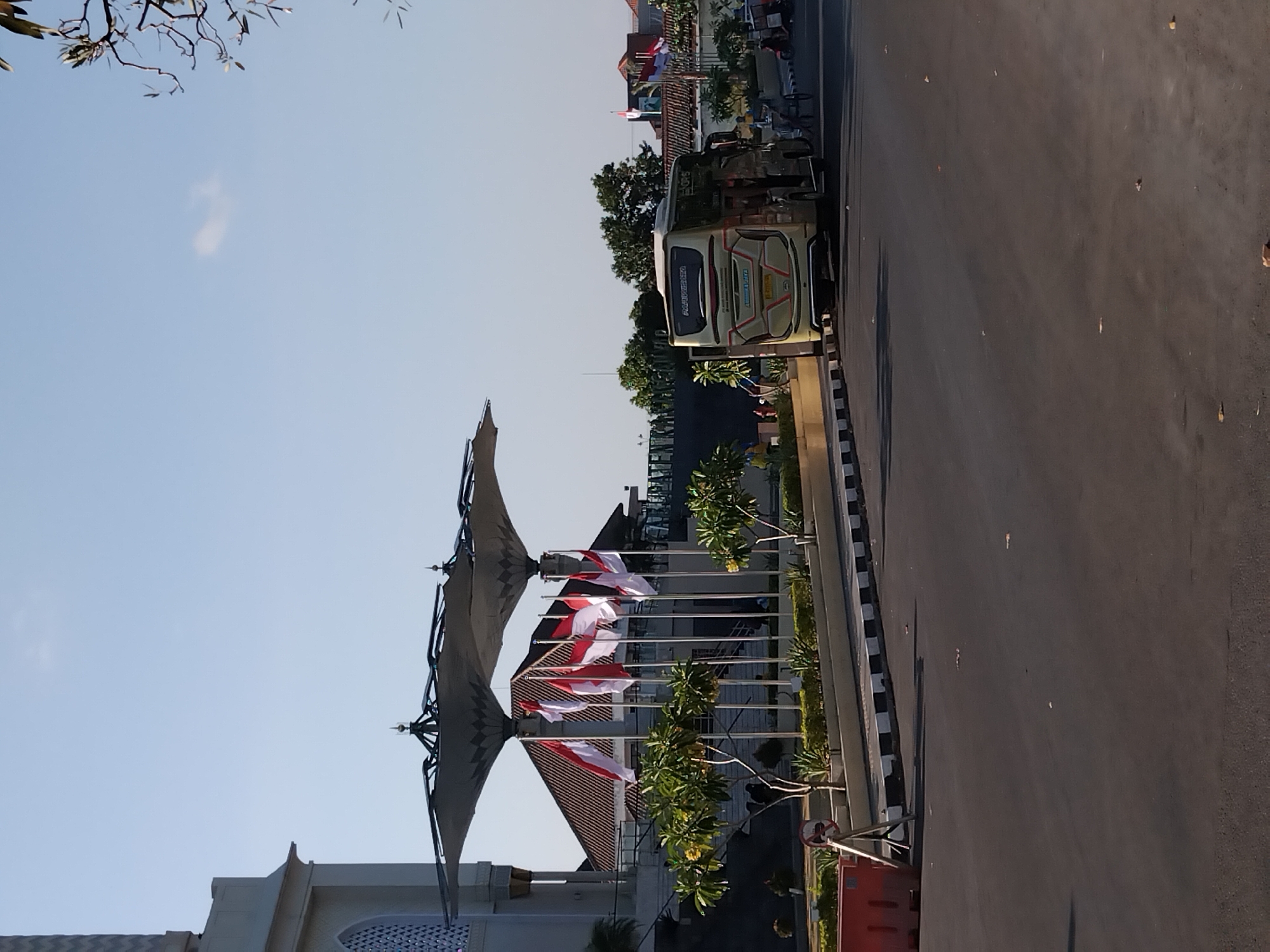 The image depicts the front entrance area of the Radya Pustaka Museum in Surakarta, Indonesia, not Karanganyar as stated. The scene is bathed in bright sunlight under a clear blue sky. Dominating the foreground is a paved road, with a distinct curb marked by alternating black and white stripes. To the left, a bright orange traffic barrier is visible, accompanied by a metal stand with a circular sign indicating prohibition.
Behind the barrier, a landscaped area features several small trees with light green foliage and scattered yellow flowers, bordered by manicured hedges. Several Indonesian flags are prominently displayed on flagpoles, situated under large, umbrella-like awnings. These awnings are supported by dark metal structures and are a notable architectural feature of the entrance.
In the background, the facade of a building, presumably part of the museum complex, is visible, characterized by its light-colored walls and an arched window with intricate lattice work. A bus, golden in color with "PAHALAWAN" written on its rear, is parked to the right of the scene, its doors open, suggesting passengers may be disembarking or embarking. Further to the right, beyond the bus, a horse-drawn cart (dokar) is partially visible, and a red motorcycle with a sidecar is parked. Trees with dark green foliage form a backdrop to the bus and the street. The overall impression is that of a public space, possibly a tourist attraction, on a sunny day.