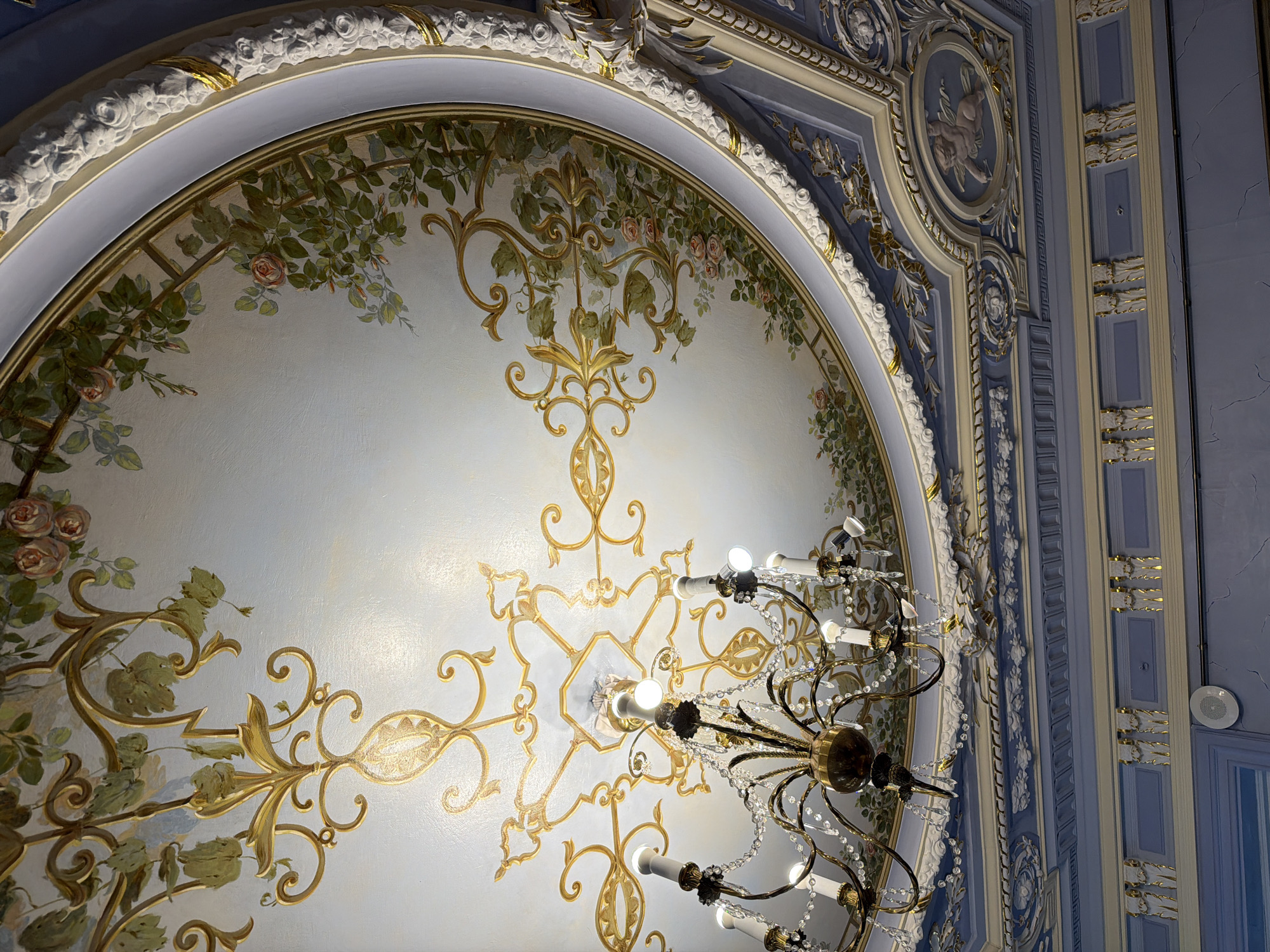 The image captures an ornate ceiling with intricate details, likely in a historical building. The ceiling is painted with a sky-blue background, adorned with delicate floral motifs of roses and vines in shades of pink, green, and gold. A gilded, Baroque-style chandelier hangs prominently in the center, featuring multiple candle-like lights and crystal embellishments. Surrounding the central painted dome is a decorative stucco molding with floral patterns and gold accents. Below this, a blue ceiling section features more elaborate molding, including a depiction of a cherub within an oval frame, along with further gilded ornamentation. The overall impression is one of opulence and historical grandeur. This ceiling is located in the Nizhny Novgorod State Academic Opera and Ballet Theatre in Nizhny Novgorod, Russia.