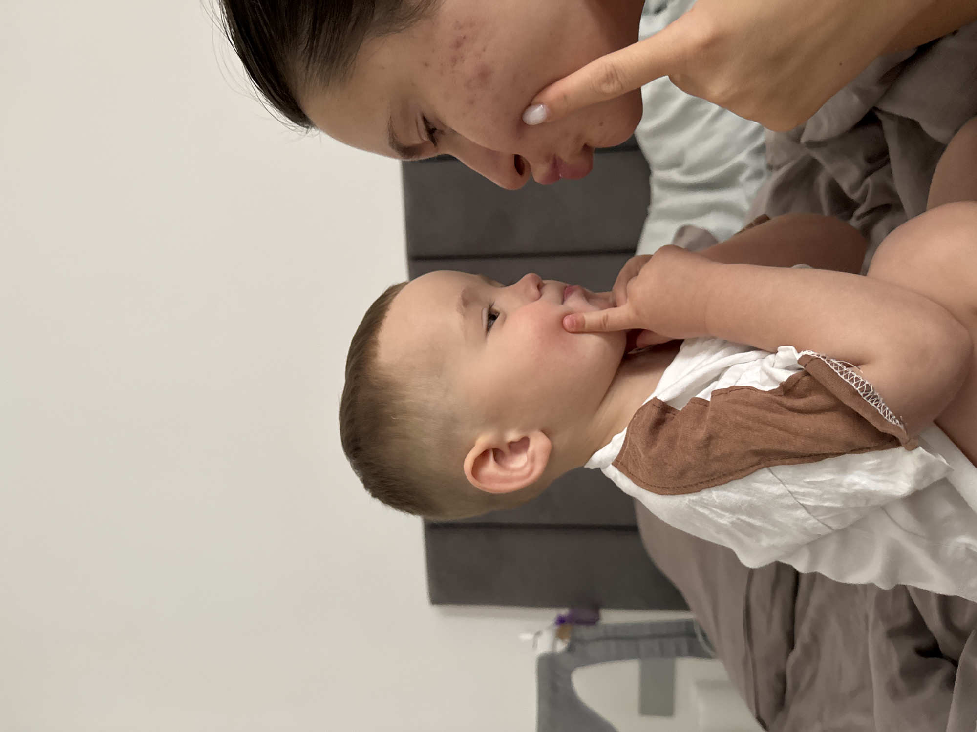 A woman and a baby are face to face, close to each other. Both are touching their faces with their fingers. The woman has dark hair pulled back from her face, and is wearing a light-colored top. The baby has light hair and is wearing a white shirt with brown sleeves. They are on a bed with gray bedding and a gray headboard. The location is Odintsovo, Russia, but the indoor setting does not provide additional geographic information.