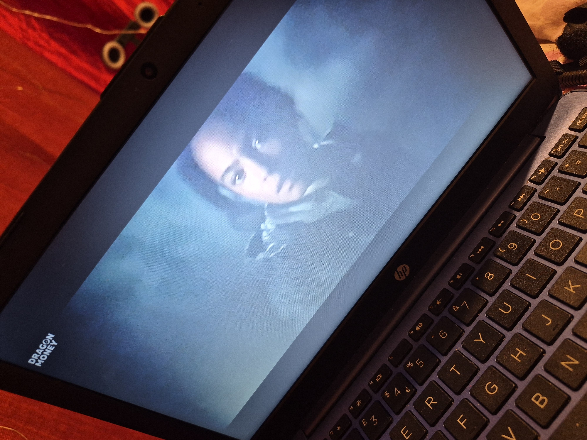 The image shows a close-up, angled view of a laptop screen displaying a scene from a movie or show. The screen shows a person's face with a concerned expression, partially obscured by a smoky or hazy effect. The top left corner of the laptop screen displays the text "DRAGON MONEY" in white letters. The laptop keyboard, primarily in a dark color with yellow lettering, is visible in the foreground, with keys like 'F', 'G', 'T', 'Y', 'B', 'H', 'N', 'J', and 'K' clearly discernible. The laptop is open, and the background is a blurry red fabric with some string lights, suggesting an indoor setting. There is no identifiable information within the image that indicates a specific location in the city of Sandwell, United Kingdom.