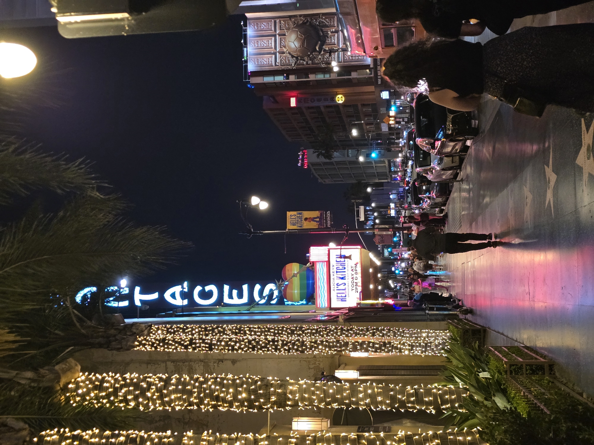 This is a vertical, eye-level, nighttime photograph of the Hollywood Walk of Fame in Los Angeles, California. The foreground features palm trees wrapped in warm white string lights. To the left, the neon sign for "PANTAGES" is visible, with individual letters illuminated in blue. Further down the sidewalk, a large sign advertises "ALICIA KEYS' HELL'S KITCHEN," with a rainbow-colored heart above it. People are walking on the sidewalk, some appearing to be tourists. Cars are parked along the street on the right side of the image, and more buildings with illuminated signs are visible in the background. The sidewalk itself is paved and reflects the ambient light. The overall scene depicts a bustling urban environment at night, characteristic of a popular tourist destination in Hollywood.