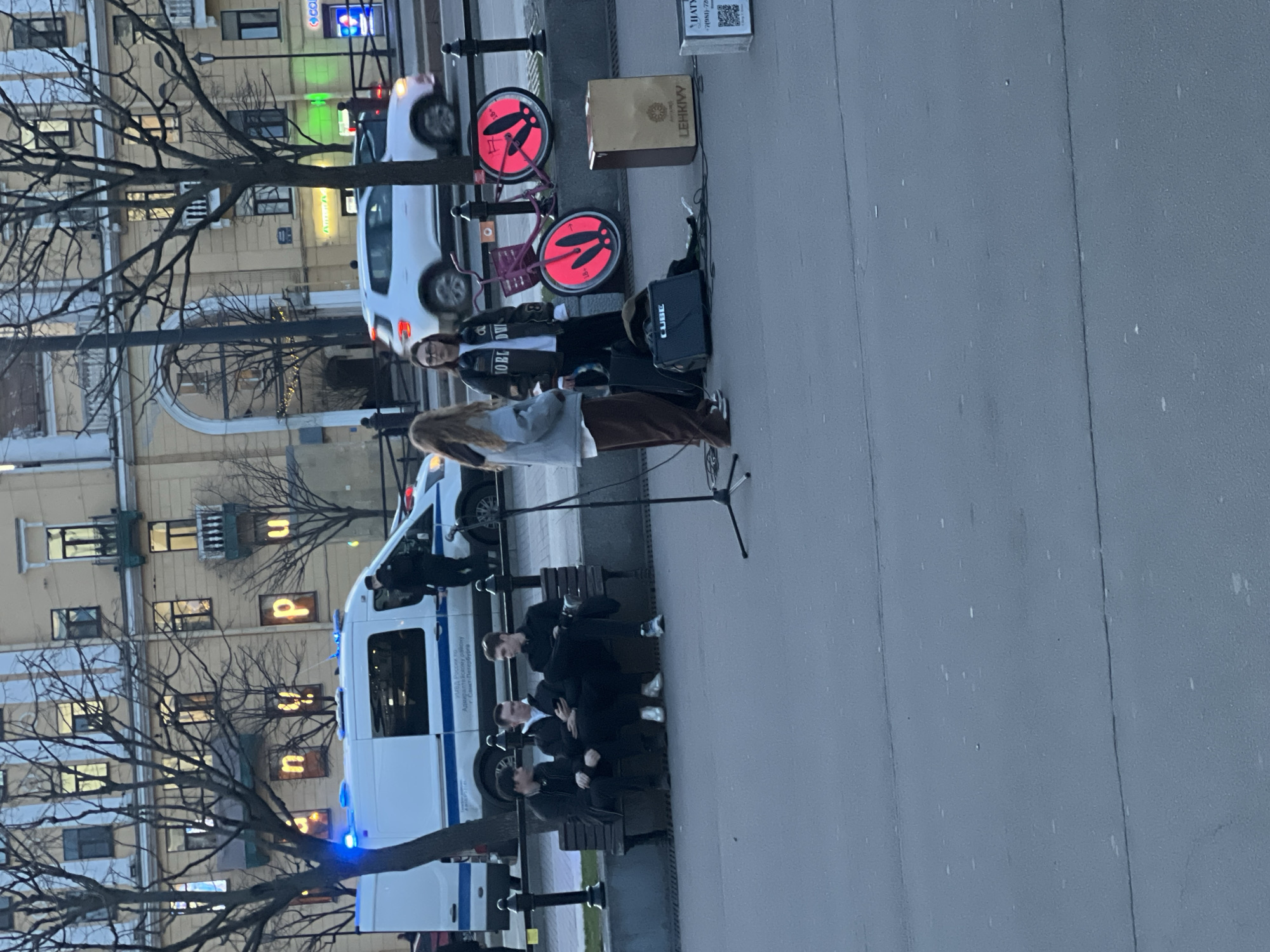 The image depicts a street scene in Saint Petersburg, Russia.
A group of individuals, presumably men, is seated on park benches near an ambulance or police van. Another group of people stands near what appears to be a busking setup with a microphone, amplifier ("CUBE"), and a cardboard box labeled "LEHKIVY." Two bicycles with bright red rims rest next to the busking area.
Several buildings are visible in the background, including one with illuminated Cyrillic script. The text on the ambulance identifies it as belonging to the Admiralty District of the Saint Petersburg Ministry of Internal Affairs of Russia (УМВД России по Адмиралтейскому району Санкт-Петербурга).