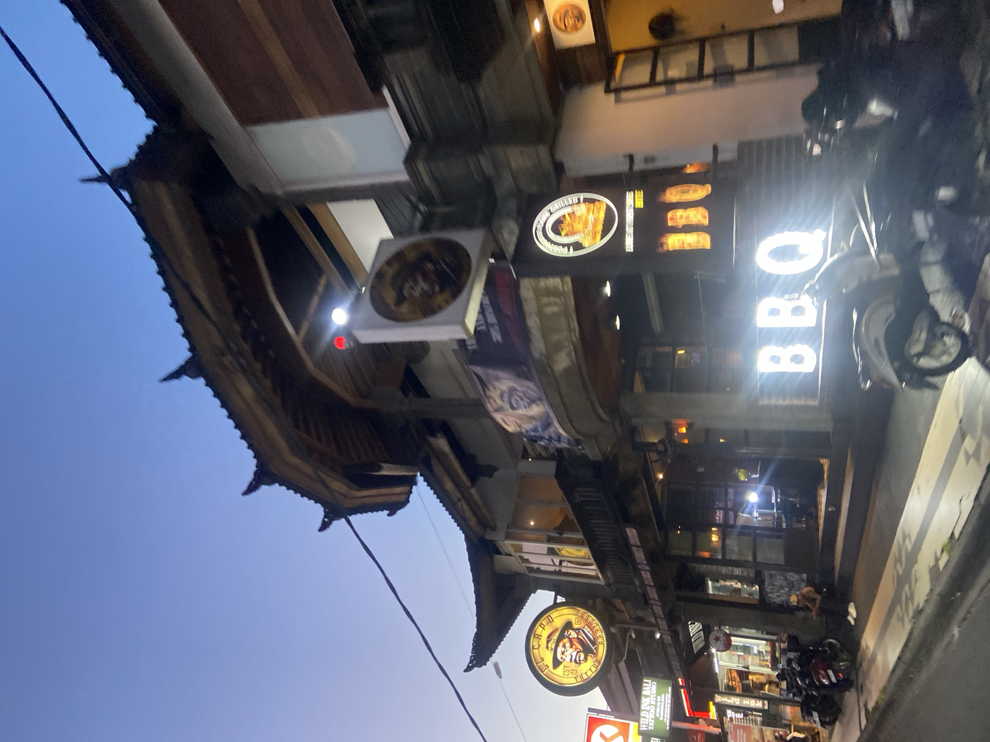This image captures a vibrant street scene at dusk or early night in what appears to be a commercial area, consistent with Kuta, Indonesia. The sky on the left side of the frame displays a deep twilight blue, suggesting the transition from day to night.
Buildings line the right side of the street, showcasing varied architecture, including structures with traditional Southeast Asian-style tiered roofs, particularly prominent on the dark-wooded building towards the foreground.
The scene is illuminated by numerous glowing signs advertising different businesses. A large, vertically oriented sign prominently displays "BBQ" in bright white neon letters. Closer to the viewer, a circular sign features a cartoonish image of a man in a hat, surrounded by the text "WILD INK BALI TATTOO STUDIO". Another circular sign further down advertises "GRILLED", with additional text like "AUTHENTIC FLAME PORK" and an image of food. Several smaller, less distinct signs also contribute to the overall glow of the street.
Motorcycles are visible parked along the roadside or on the sidewalk, suggesting typical street activity. The combination of the dark blue sky and the illuminated storefronts creates a lively, bustling atmosphere typical of a tourist or commercial district in the evening. No specific individuals are clearly visible engaged in distinct activities.