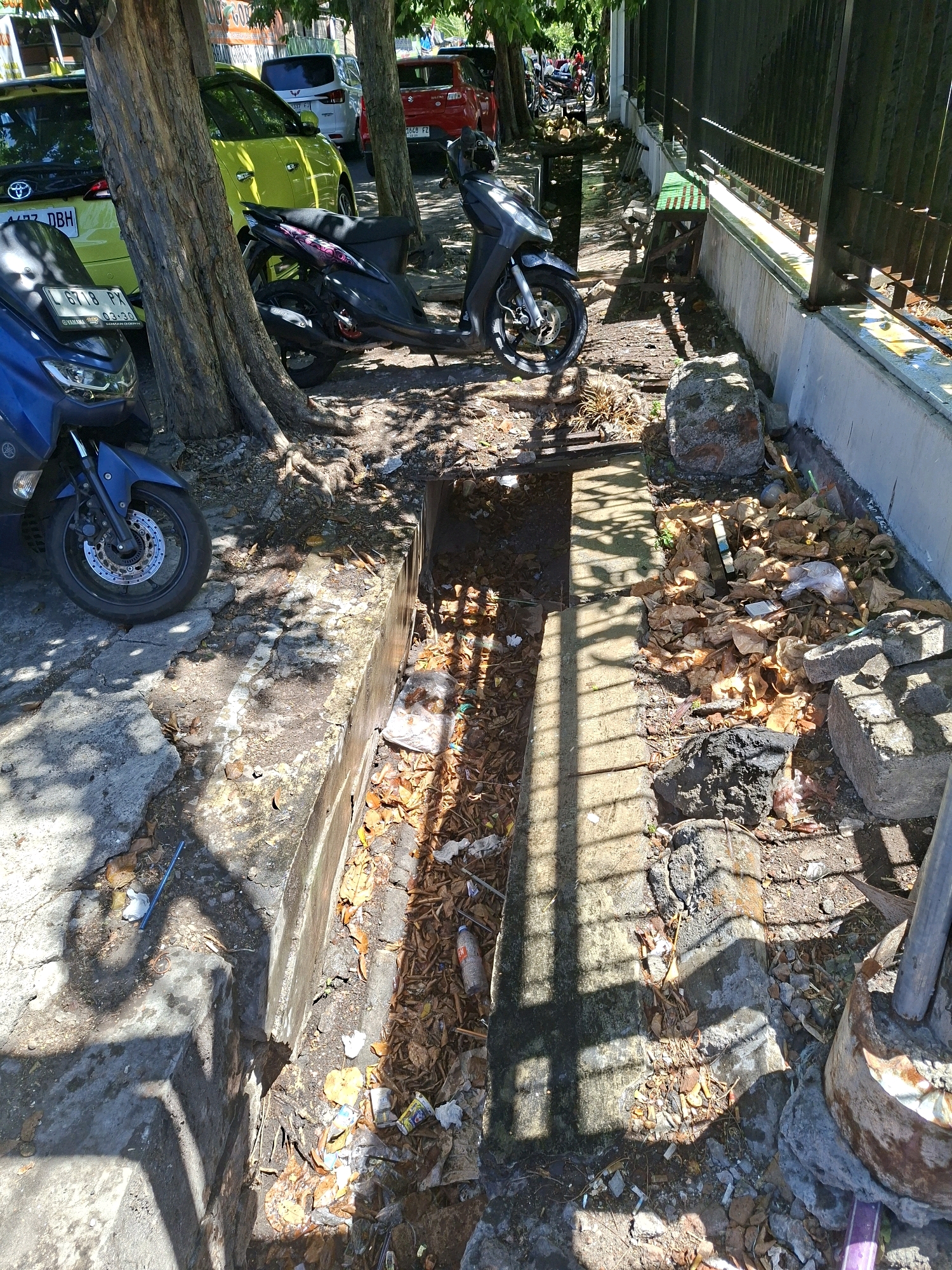 The image shows a street scene in Surabaya, Indonesia, with a focus on a drainage ditch. The ditch appears to be partially covered and filled with debris, including leaves, plastic bags, and other refuse. Several motorcycles are parked along the side of the street, suggesting a public area or a thoroughfare. The surrounding environment includes trees providing shade, a sidewalk, and a metal fence. The presence of the ditch and the debris indicate a potential issue with street cleaning or infrastructure maintenance. The lighting suggests it is daytime, with strong shadows cast by the trees and fence.