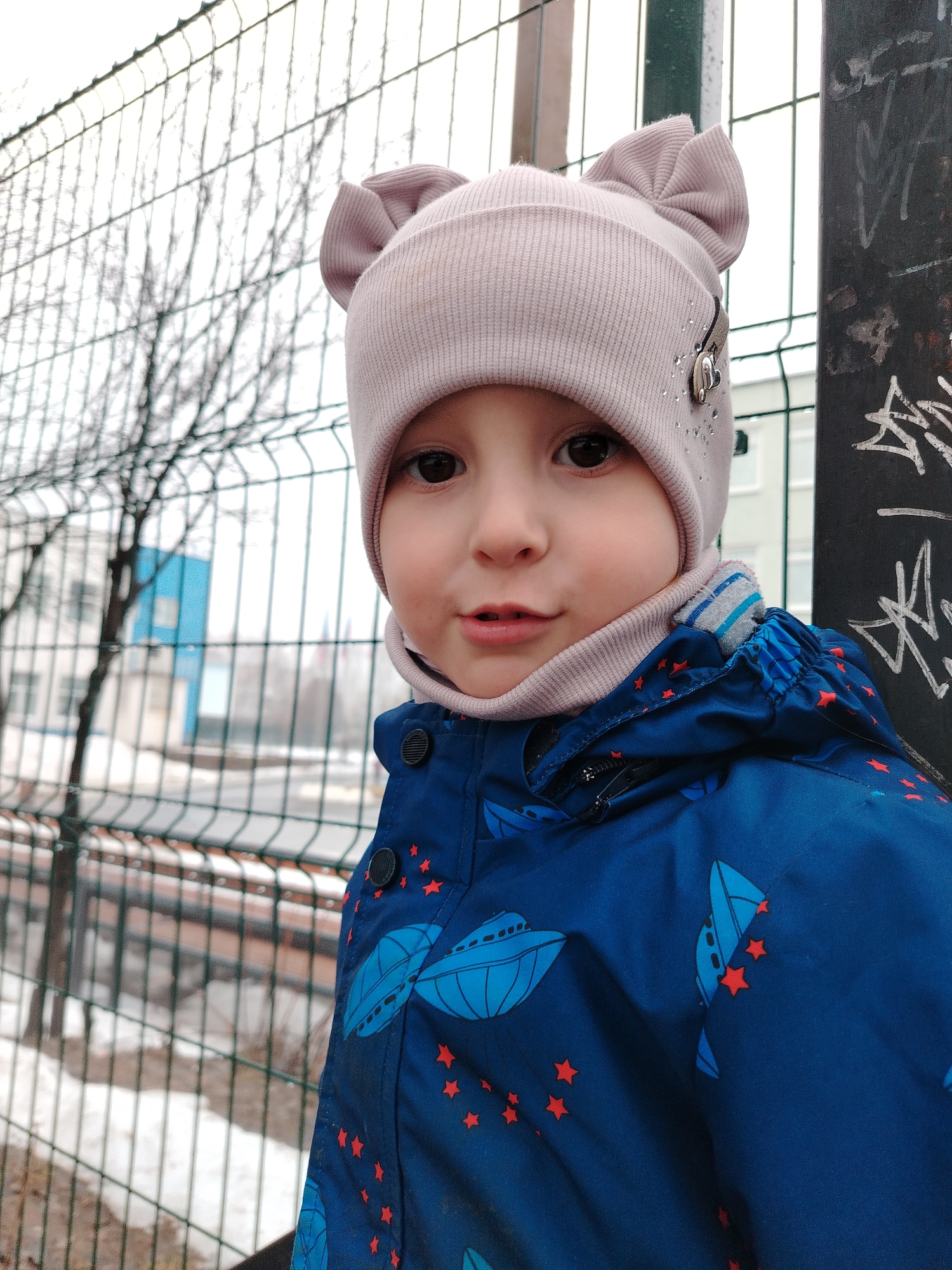 A young child, likely between 3-5 years old, stands outdoors next to a metal fence. The child is wearing a light pink hat with ear-like protrusions, a matching neck gaiter, and a blue jacket adorned with spaceship and star patterns. The child is looking directly at the camera. The background includes a metal fence, bare tree branches, a building with a blue facade, and a metal pole covered in graffiti.
Given the information, the location is the city of Chaikovsky, Russia.
