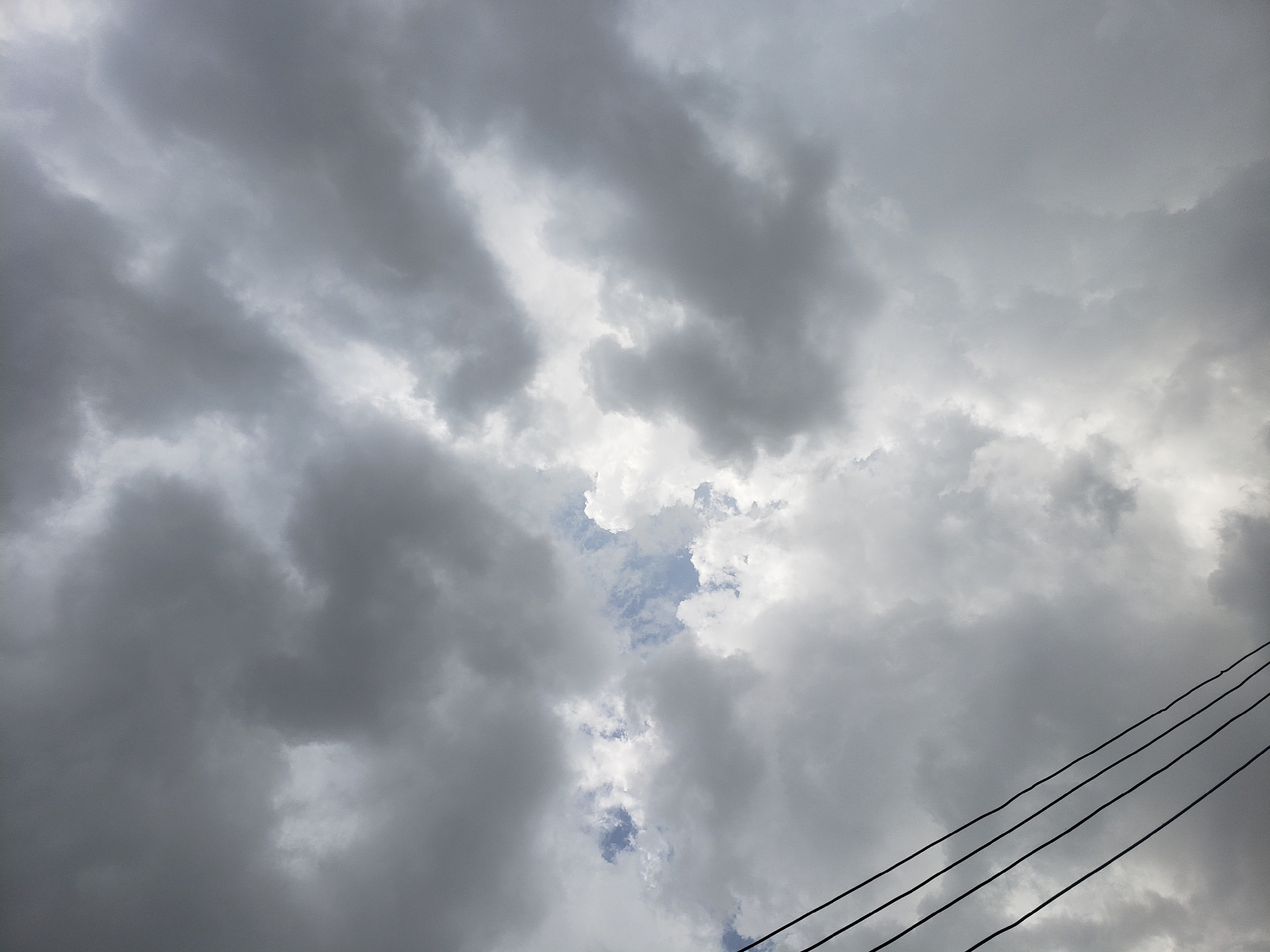 The image displays a heavily overcast sky, dominated by dense, dark grey clouds that fill most of the frame. The clouds appear thick and turbulent, suggesting a large weather system or impending rain. In the central area, a small, irregular patch of lighter cloud and glimpses of blue sky are visible, offering a break in the uniform grey. Towards the lower right corner, three parallel, dark power lines stretch diagonally across the sky, adding a man-made element to the natural scene. The overall atmosphere conveyed is one of a dramatic and possibly somber day. There are no people, other objects, or discernible activities taking place. No text is visible. The scene is set in Adankolo, Nigeria, under what appears to be pre-storm or heavily clouded weather conditions.