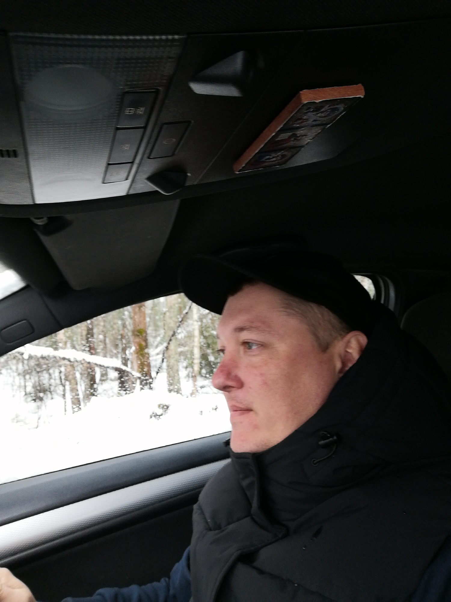 A close-up, eye-level shot captures a person inside a car, looking out at a snowy landscape. The subject, a person with short light-colored hair and a fair complexion, is seen in profile, facing right. They are wearing a dark baseball cap, a black puffy vest over a dark long-sleeved shirt, and have the collar of a jacket pulled up around their neck. Their expression appears neutral, focused on the view outside the vehicle.
The setting is inside a car, with elements of the car's interior clearly visible. The upper part of the image shows the car's ceiling, including an overhead console with several buttons. Some unidentifiable symbols or characters are faintly visible on these buttons. A small, rectangular item with a wooden-like frame and images (possibly stickers or decorations) is attached near the console, hanging downwards. A sun visor is visible above the window. The car's door panel and window frame are visible, featuring a textured trim.
Outside the car, a winter scene unfolds. Snow-covered trees and possibly snow on the ground are visible through the window, indicating cold, snowy weather. The light suggests it is daytime. The trees appear to be part of a forest or wooded area. No specific geographical cues are discernible. The person's hand is partially visible in the lower left, appearing to rest near the steering wheel or door armrest.