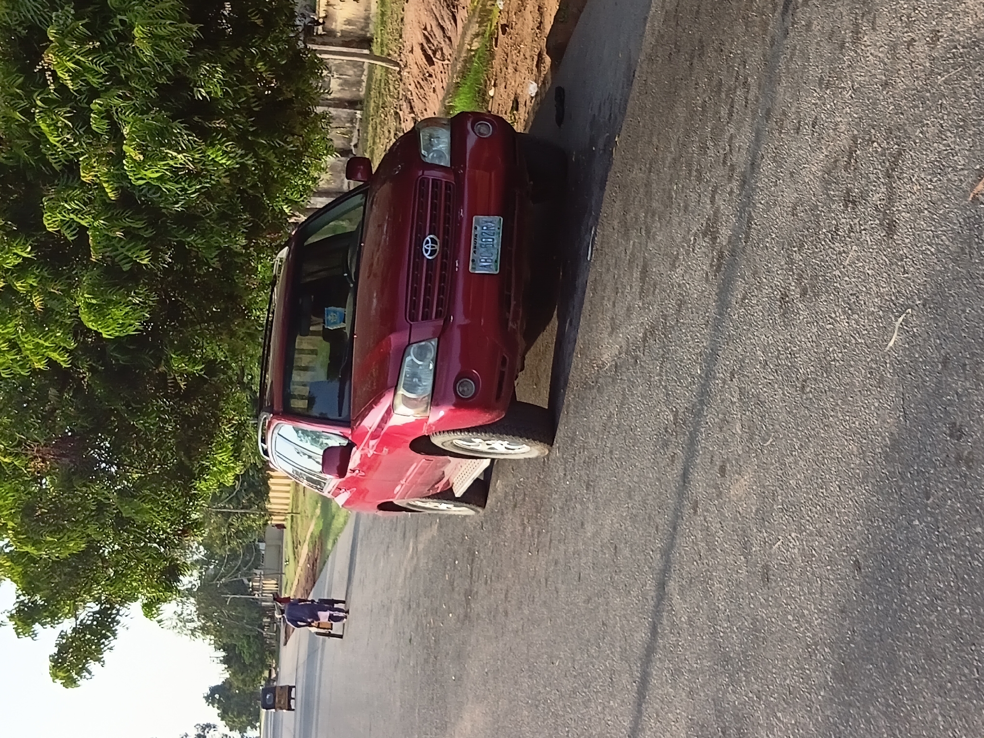 A red Toyota Highlander SUV is parked on the right side of a paved road, casting a shadow on the asphalt. The SUV is facing towards the viewer, and its license plate reads "ABC 102RX". A person in a dark blue robe is walking on the left side of the road, pushing a cart. In the distance, a yellow three-wheeled vehicle is driving away from the viewer. The road is lined with trees and a concrete wall. The sky is overcast. The scene appears to be in Jalingo, Nigeria.