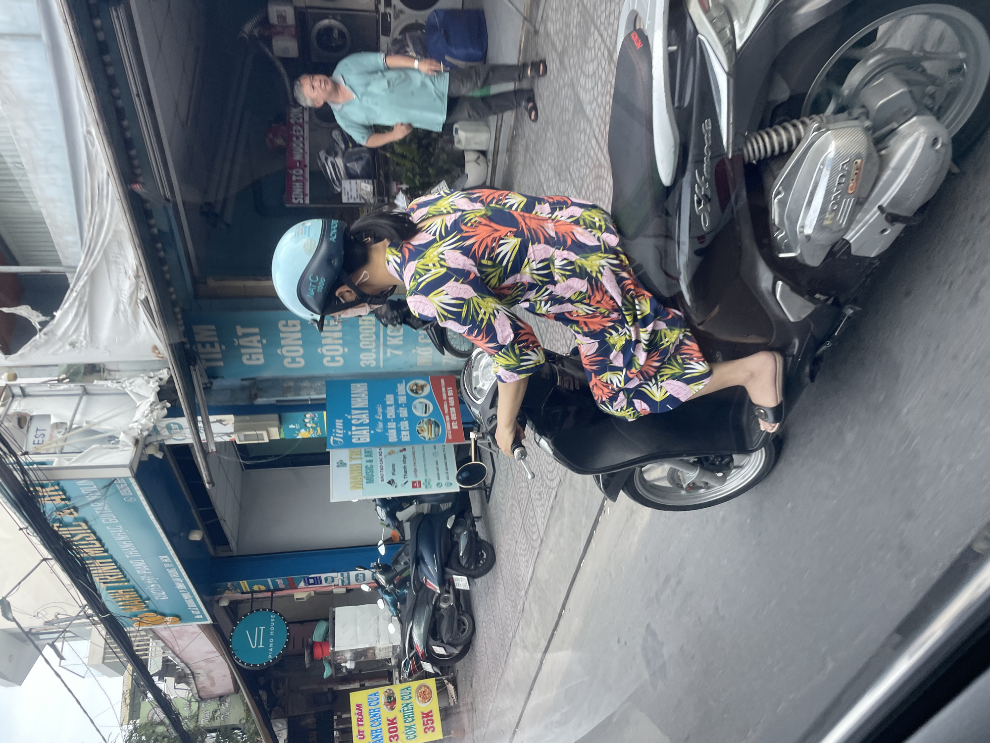 A woman wearing a colorful floral dress and a light blue helmet is riding a dark-colored scooter on a street in Thủ Đức, Vietnam. She is looking to the right, away from the camera. In the background, there is a row of businesses, including a music and art store with signs in Vietnamese, and a laundromat with washing machines visible. A man in a light blue shirt and dark pants is standing outside the laundromat. Several other scooters are parked on the sidewalk. The street appears to be a busy urban area with overhead power lines.