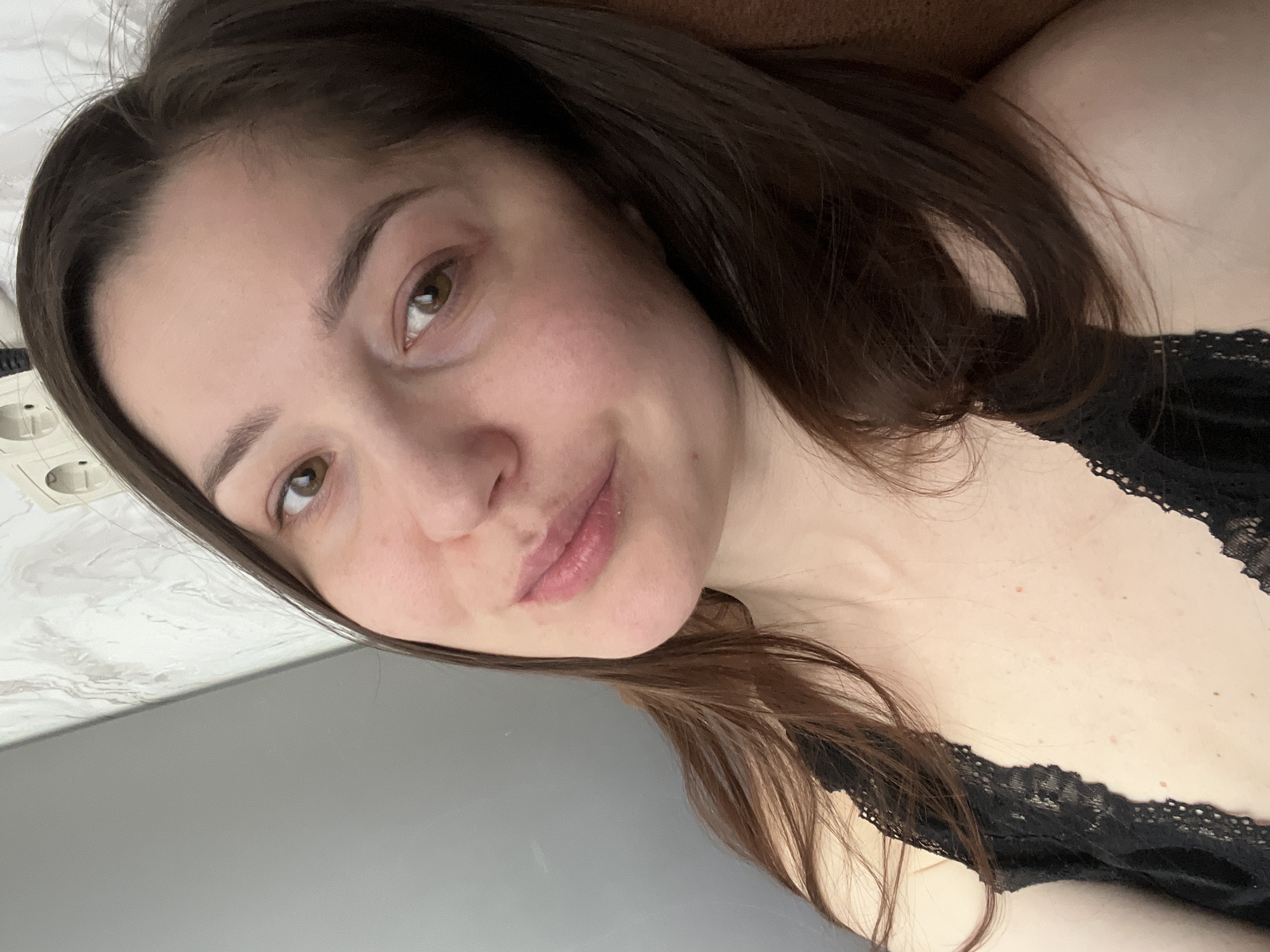 A close-up selfie depicts a woman with fair skin and brown hair. She is lying down, with her head tilted towards the left of the frame. The woman has brown eyes, and her lips are slightly parted. She is wearing a black lace top. The background includes a gray surface on the left and an electrical outlet on the top left. The location is Unknown, Unknown.