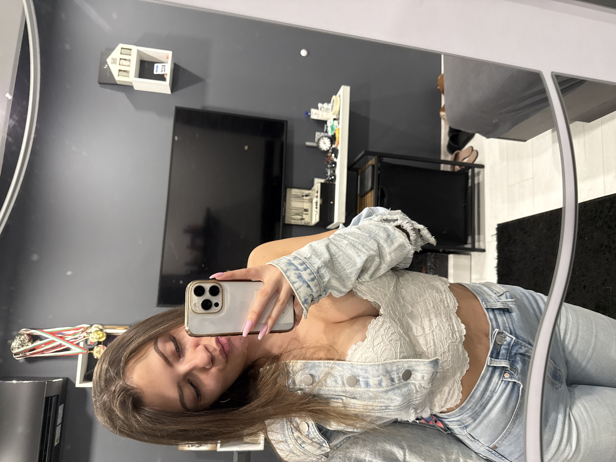 A woman takes a selfie in a mirror. The background features a dark gray wall, a television, a white decorative house on the wall, and a display of medals. Behind a black shelf, a person wearing brown shoes is visible in the mirror's reflection. The location appears to be indoors, likely a private residence. There is no evidence to suggest a specific location in Sofia, Bulgaria.