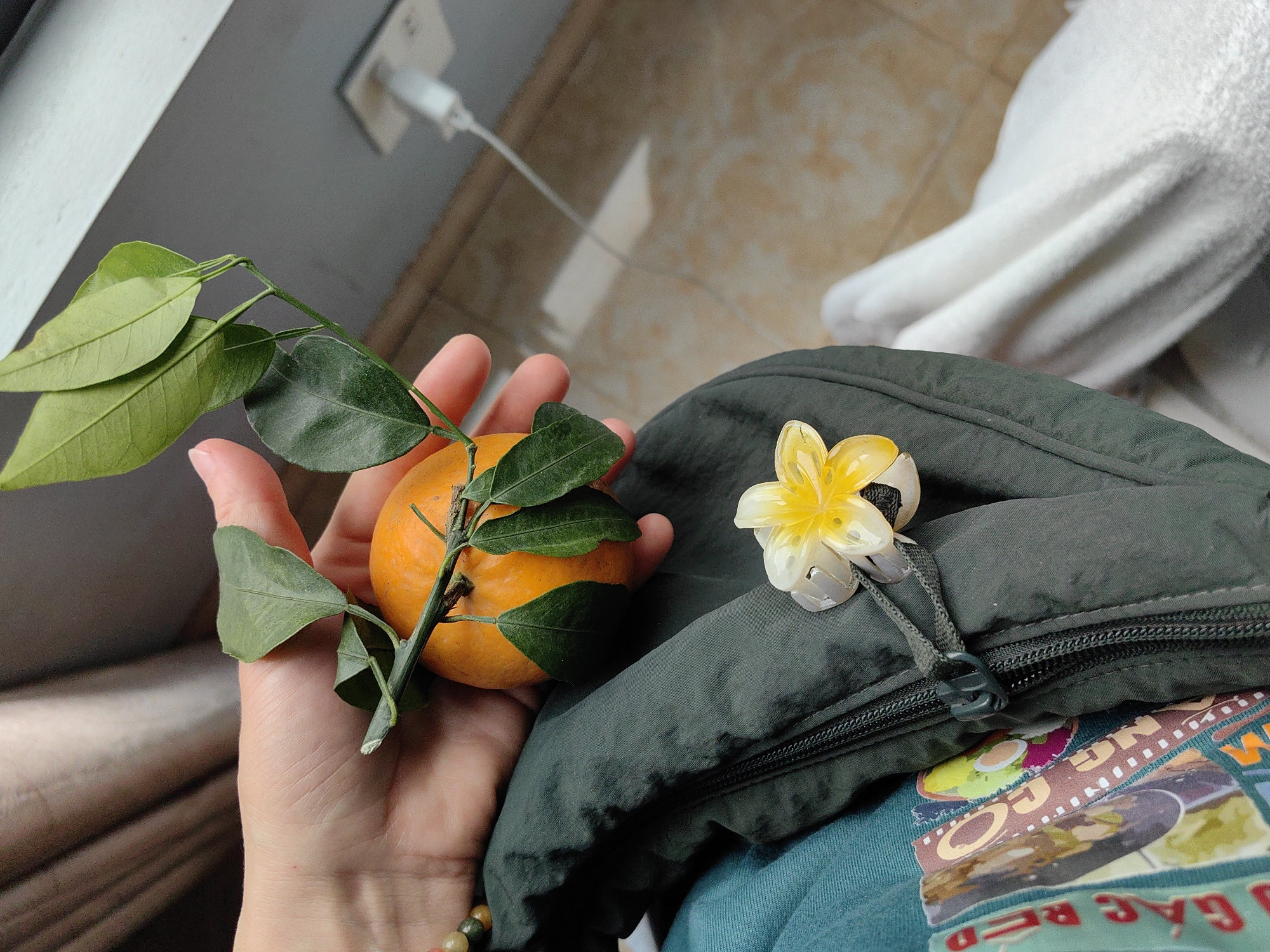 A hand holds an orange with attached stem and leaves. A decorative flower is pinned to a dark green backpack on a lap. The background includes part of a wall with an electrical outlet and a white towel. The image appears to have been taken indoors. The city of Bai Chay Ward, Vietnam, cannot be verified based solely on the image.